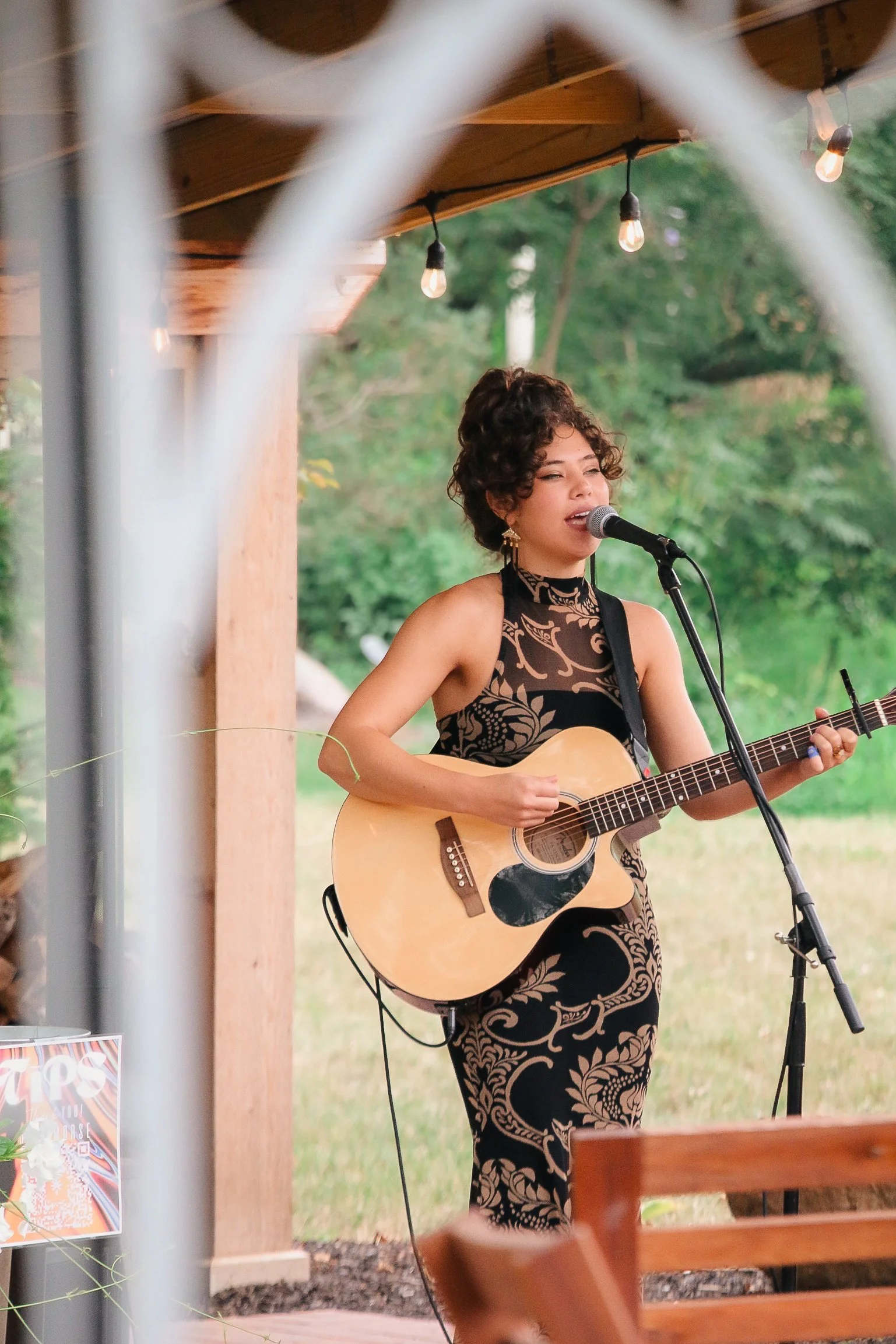 A woman with curly hair singing into a microphone and playing an acoustic guitar outdoors, wearing a black and tan intricate patterned dress.