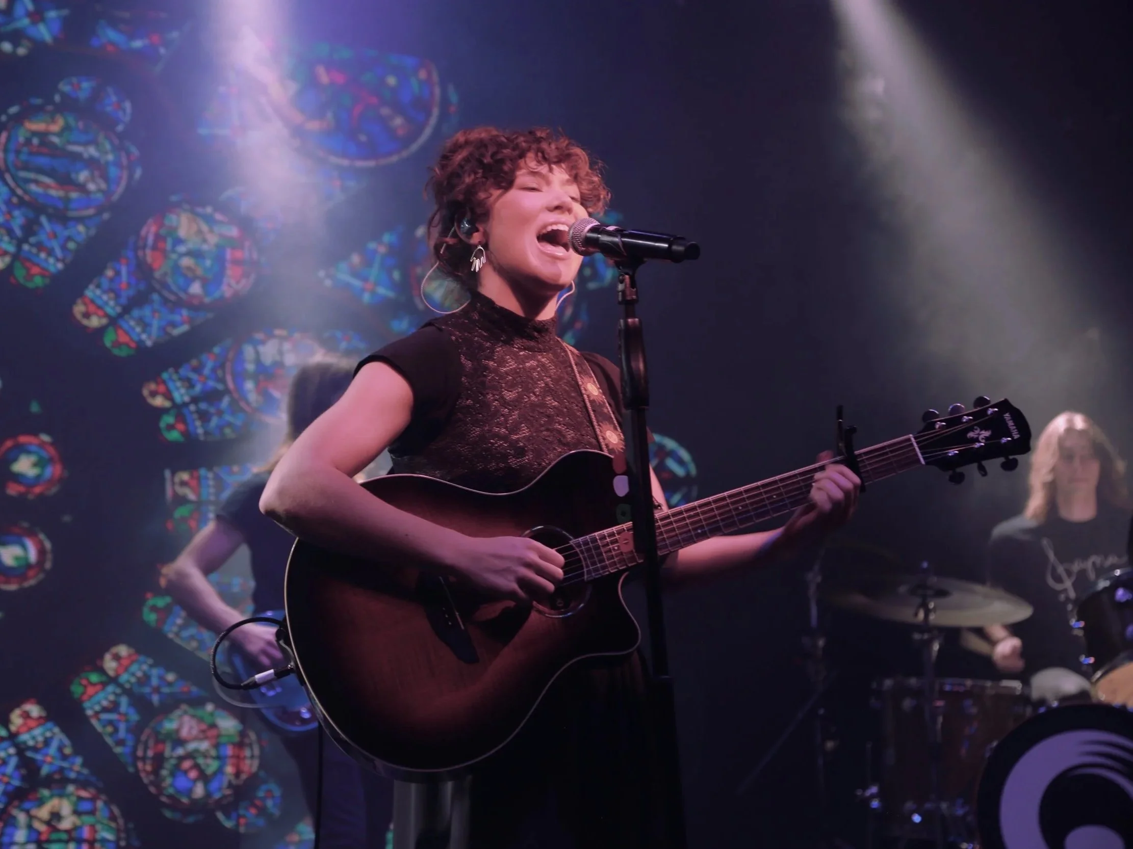 A woman with curly hair singing into a microphone while playing an acoustic guitar on stage, with colorful stained glass window artwork and stage lighting in the background.