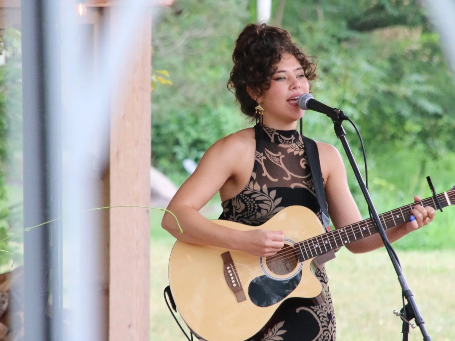 A woman with curly hair singing into a microphone and playing an acoustic guitar outdoors, with a green, leafy background.
