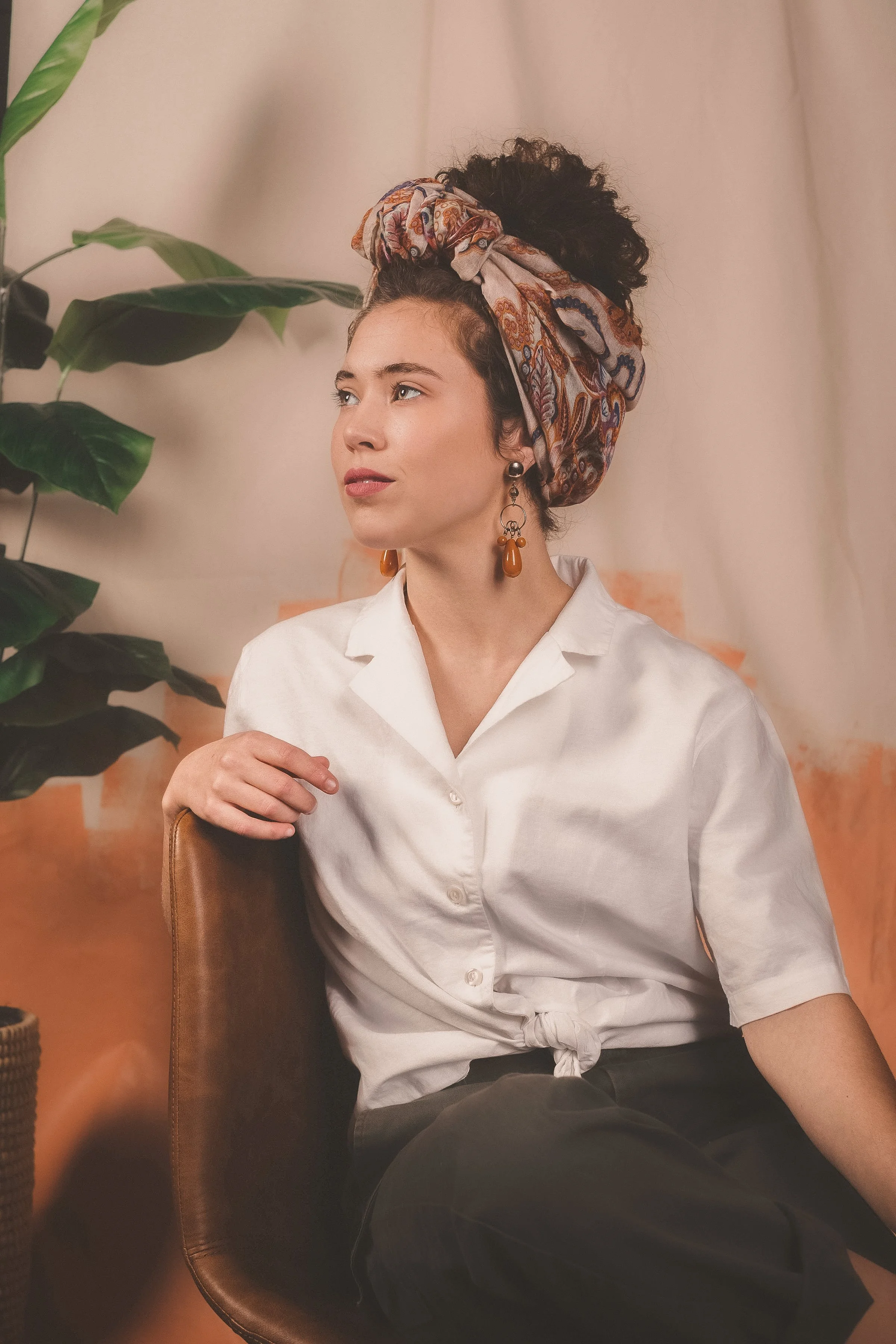 A woman sitting on a brown chair with a large green potted plant beside her. She is wearing a white shirt tied at the waist, black pants, large earrings, and a patterned headscarf. She is looking slightly to her left with a neutral expression.