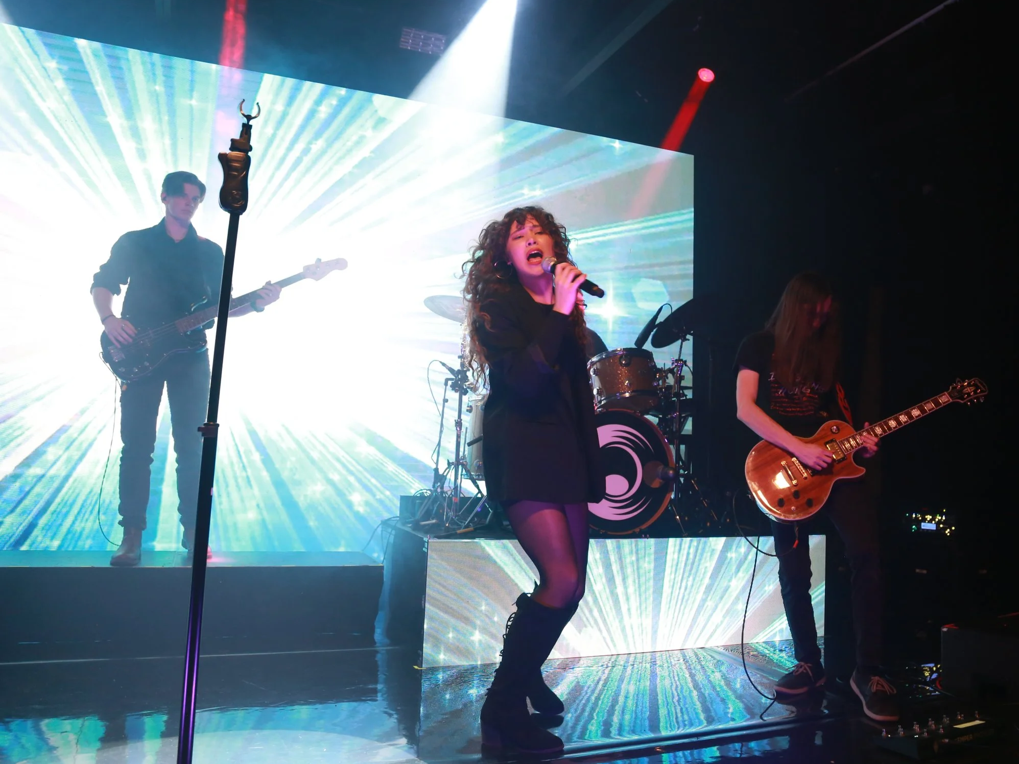 A band performs on stage with vibrant, colorful lighting and a dynamic background. A female vocalist in black clothing and fishnet stockings sings into a microphone, while male guitarists play their instruments. The scene is energetic and lively.