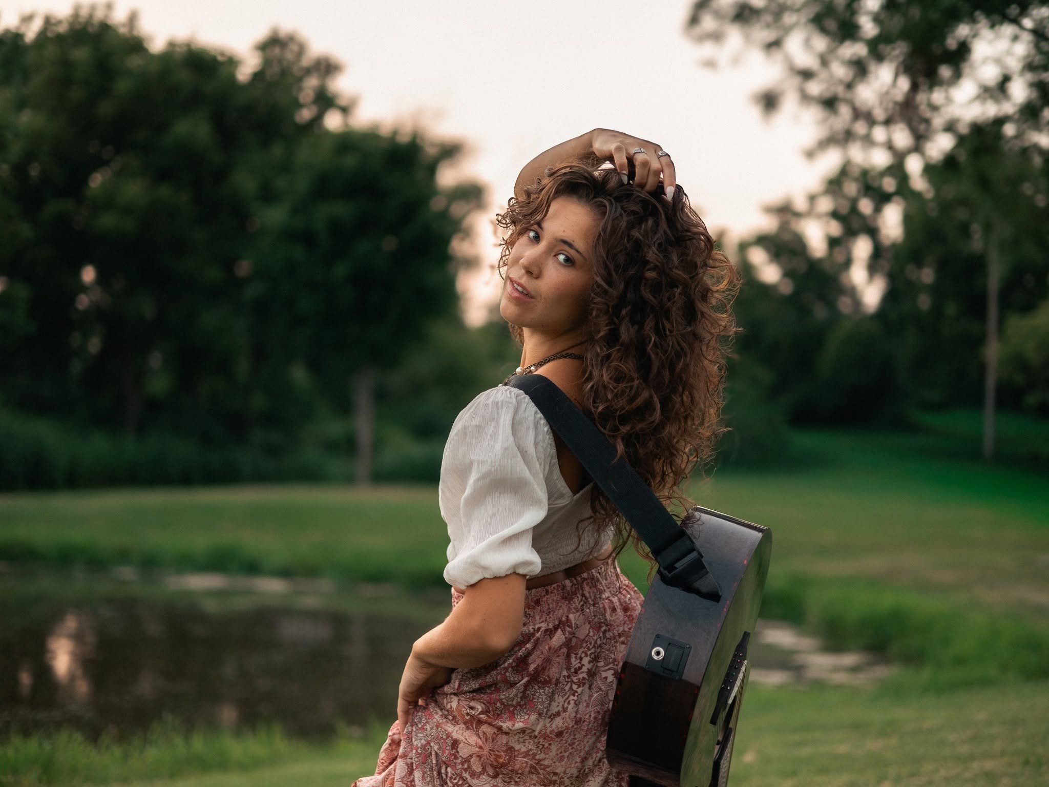 A woman with curly brown hair and light skin standing outdoors in a park, holding a guitar case and looking at the camera with a slight smile, trees and a grass field visible in the background.