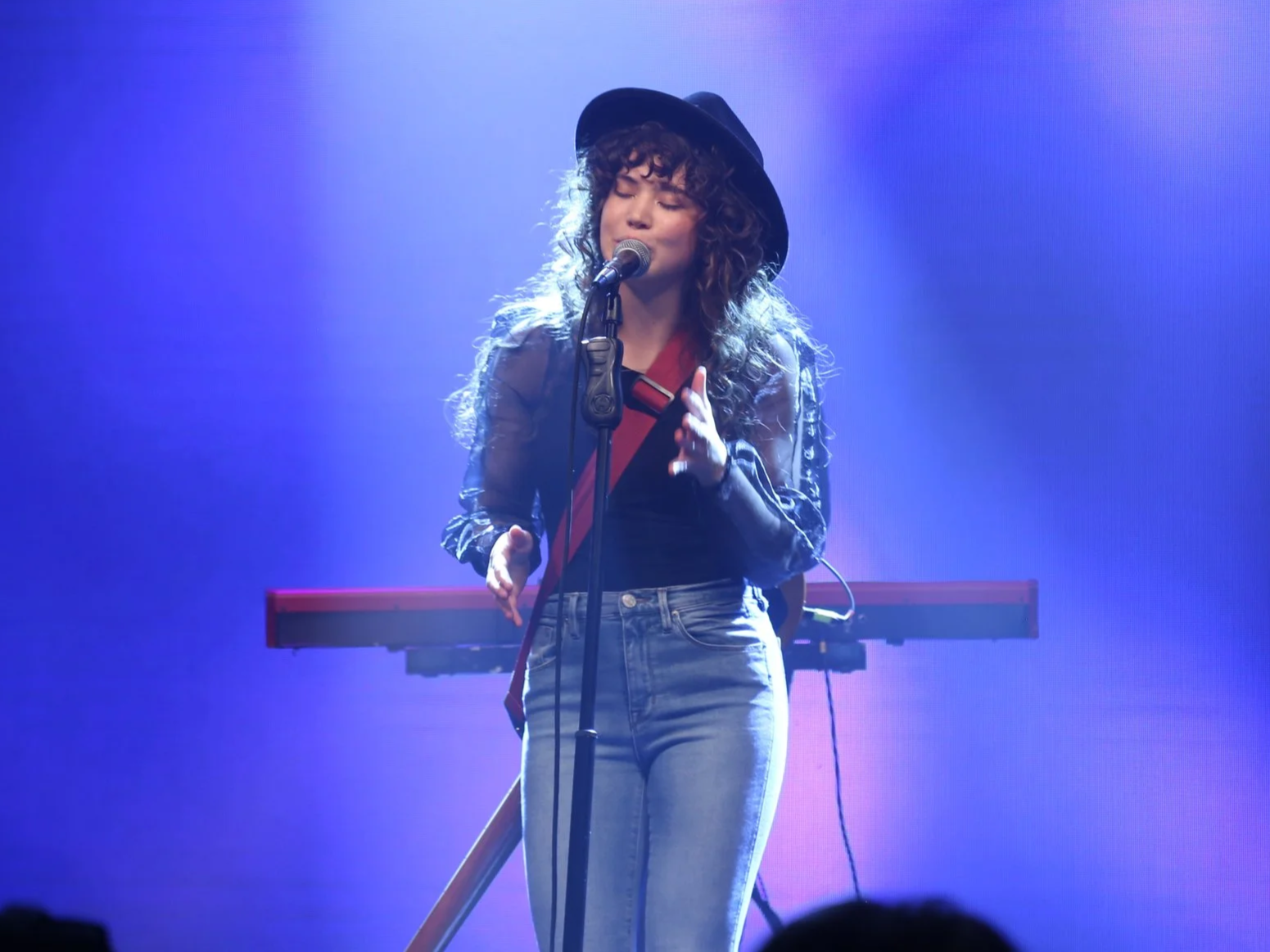 Female singer with curly hair, wearing a black wide-brimmed hat, black jacket, and jeans, singing into a microphone on stage with purple lighting and a keyboard in the background.