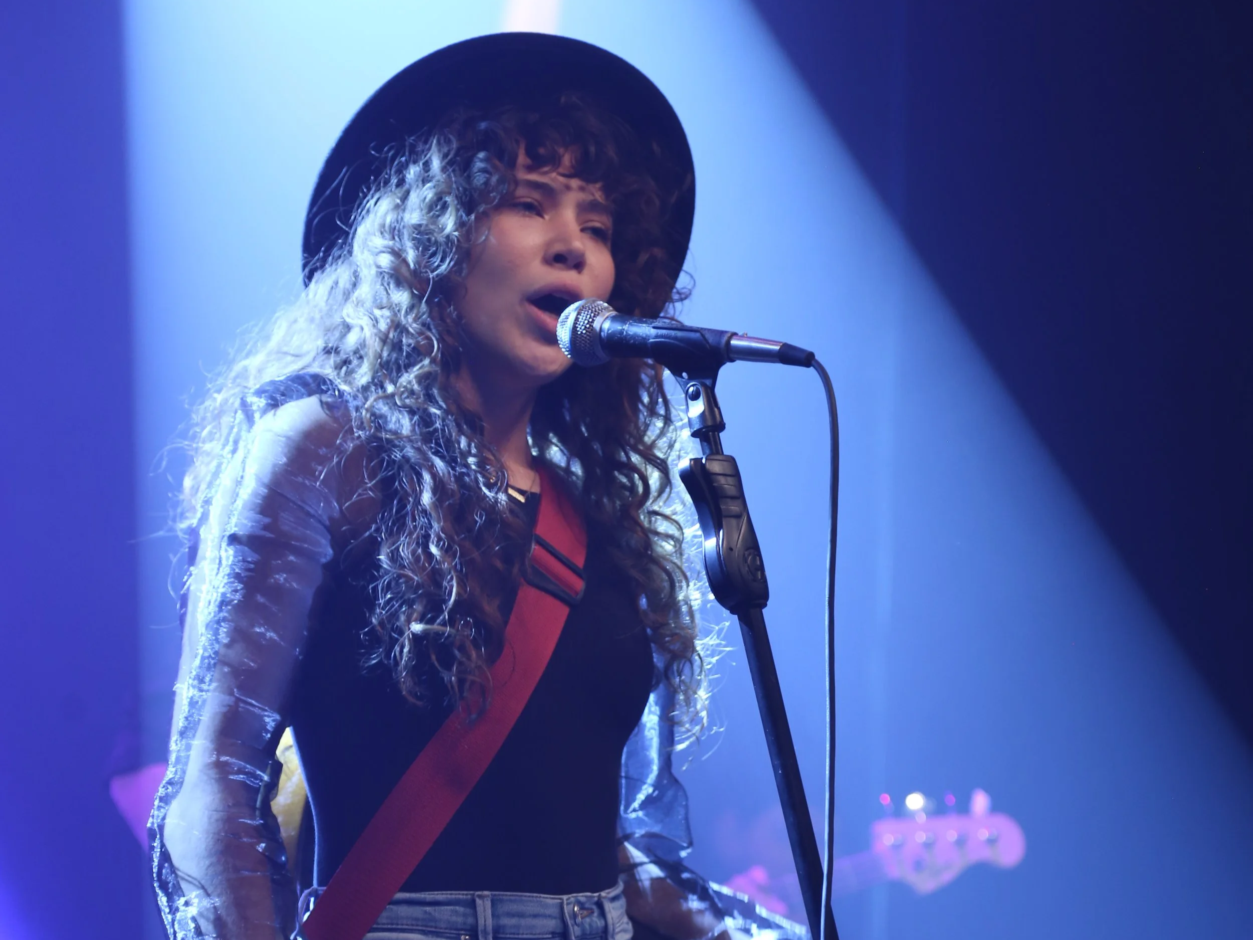 A woman singing into a microphone, wearing a black hat and a shiny jacket, in a performance setting with blue and purple stage lighting.