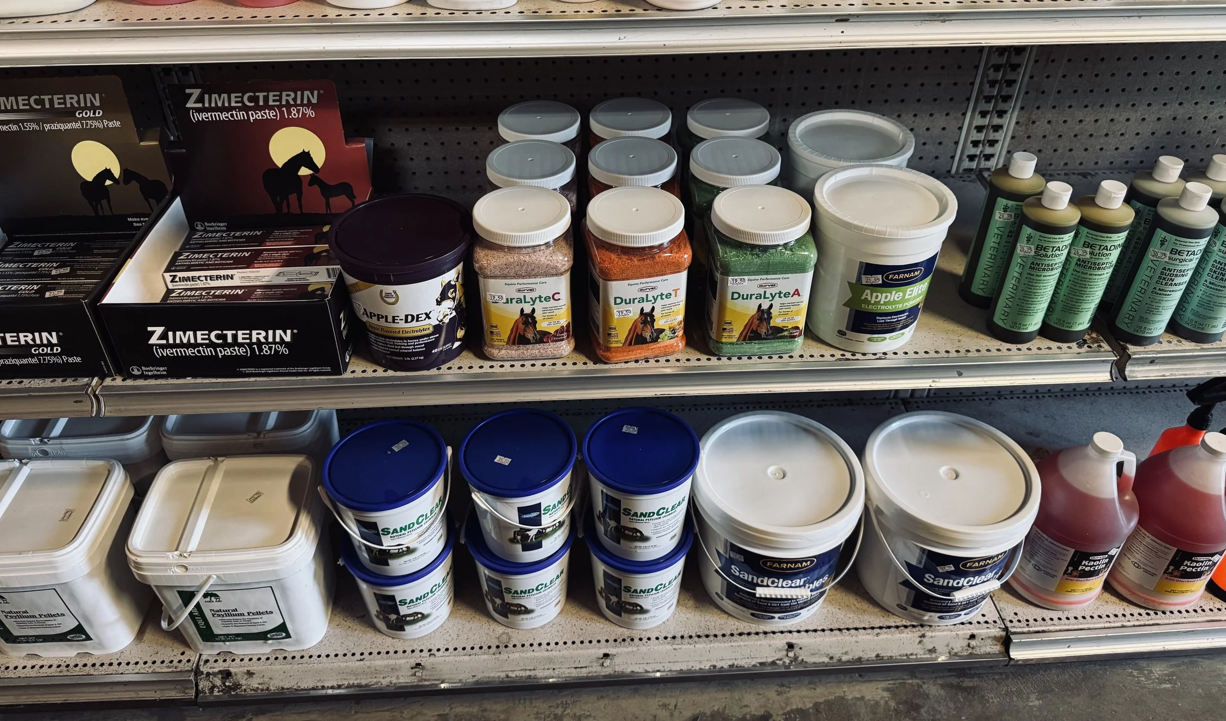 Store shelf displaying various pet medications and supplements, including wormer paste, electrolytes, and other veterinary products, some in jars and bottles.