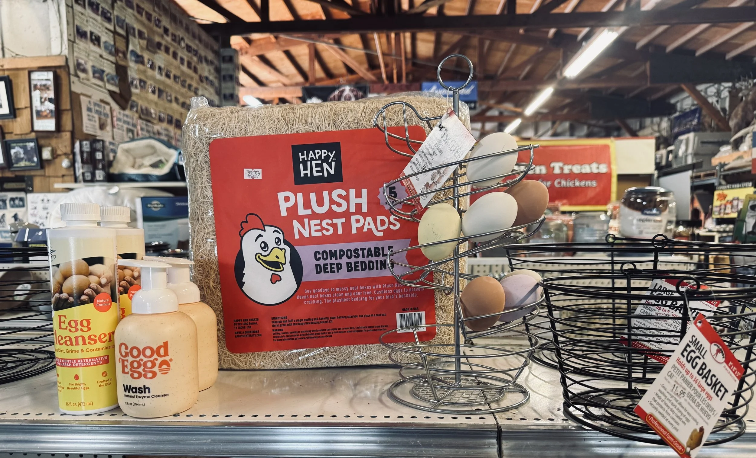 A store aisle displaying chicken egg products, including a bag of Plush Nest Pads from Happy Hen and bottles of egg wash and degreaser. The background shows a rustic store interior with wooden beams and shelves filled with various products.