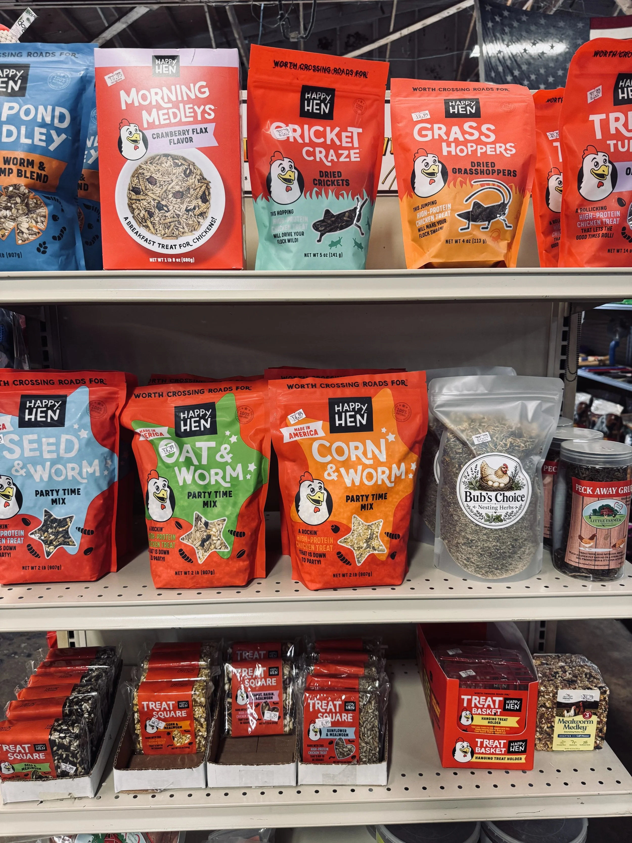 Shelf with various bags of chicken treats and snacks for birds, including dried crickets, grasshoppers, oat & worm mix, corn & worm mix, and treat squares, all branded Happy Hen.