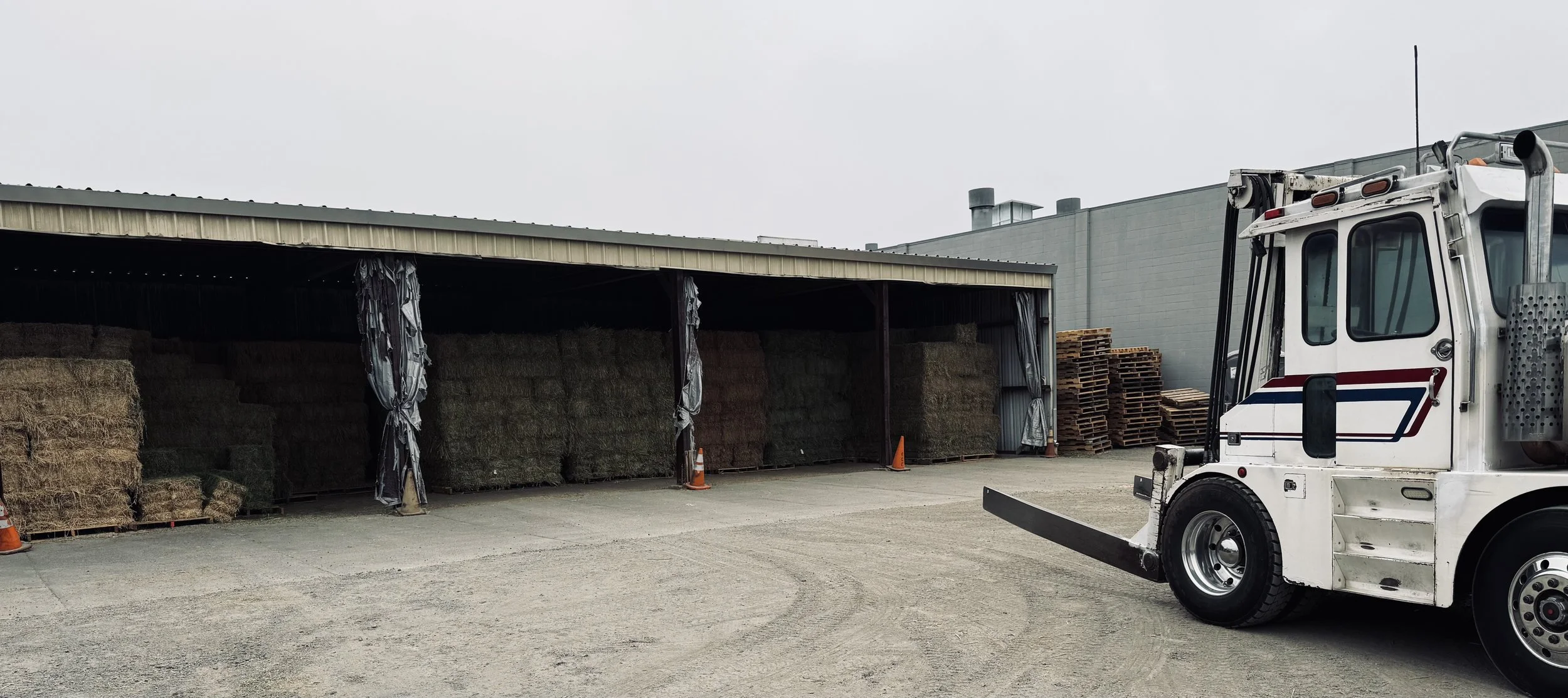 An outdoor storage area with stacks of hay bales under a metal shed and wooden pallets beside a white forklift.