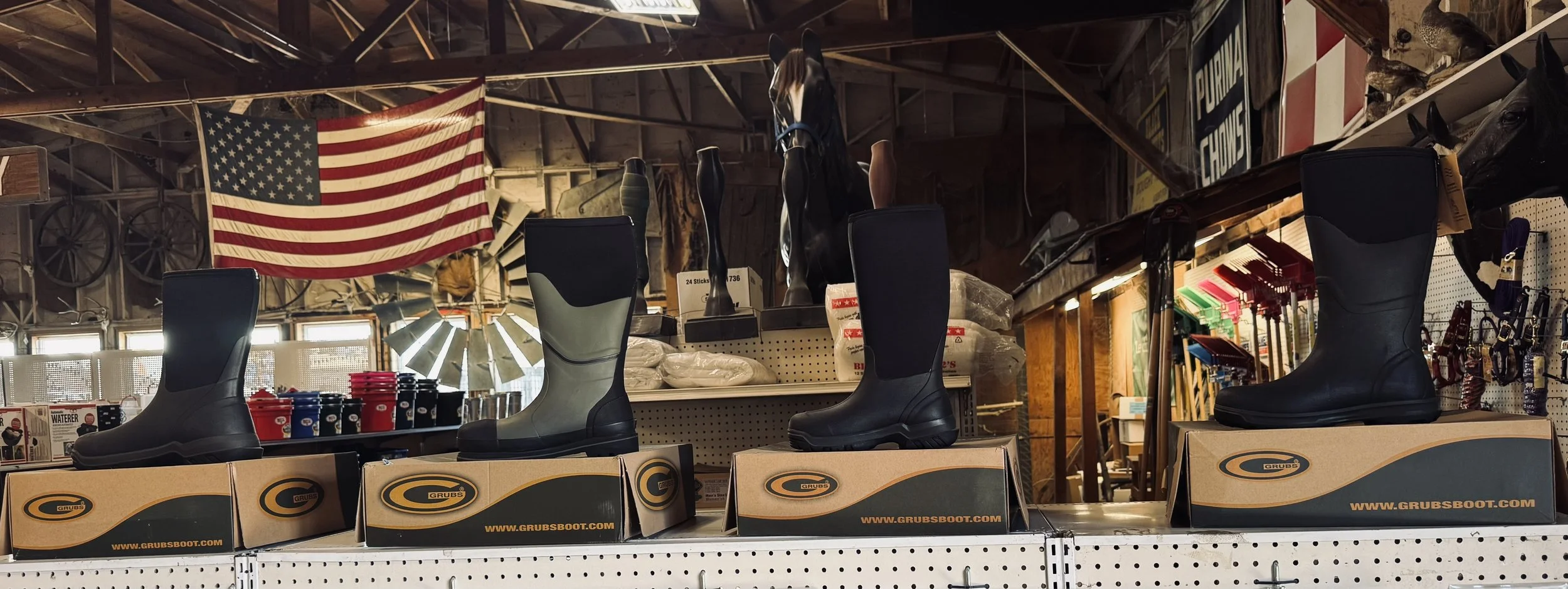 Display of rubber work boots on shelves inside a hardware store, with an American flag hanging on the wall in the background.