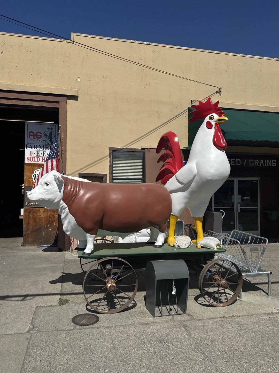A large sculpture of a cow and a rooster on a cart outside a building, with an American flag hanging nearby.