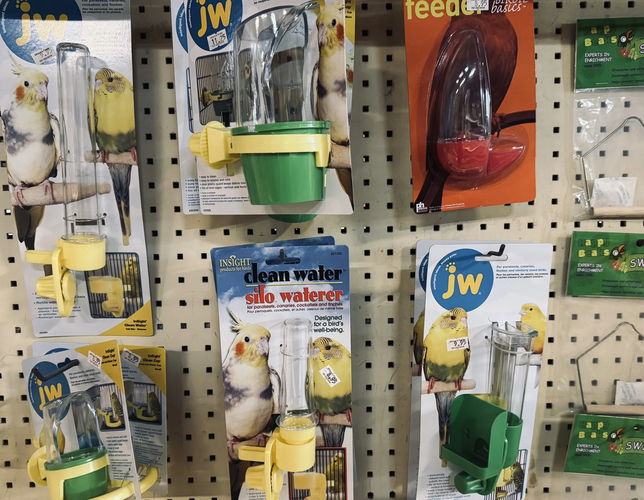 Bird watering cups and bird cage cleaning products displayed on store pegboard with bird images on packaging.