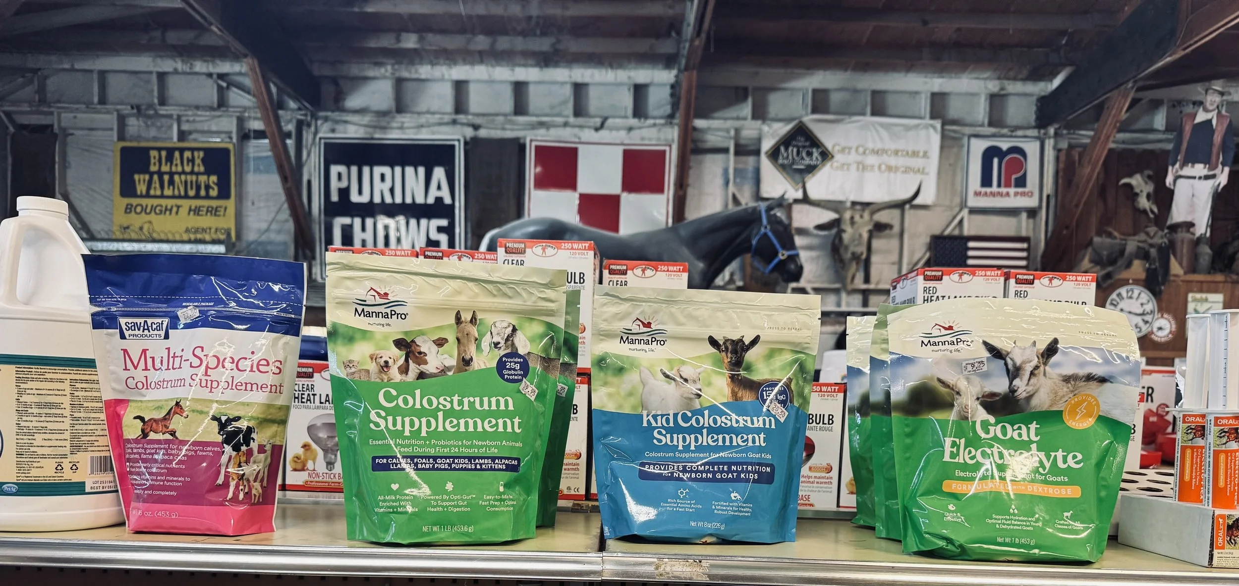 Pet supplement and food bags on store shelf, including colostrum, goat electrolytes, and multi-species colostrum supplements, with animal-themed packaging.