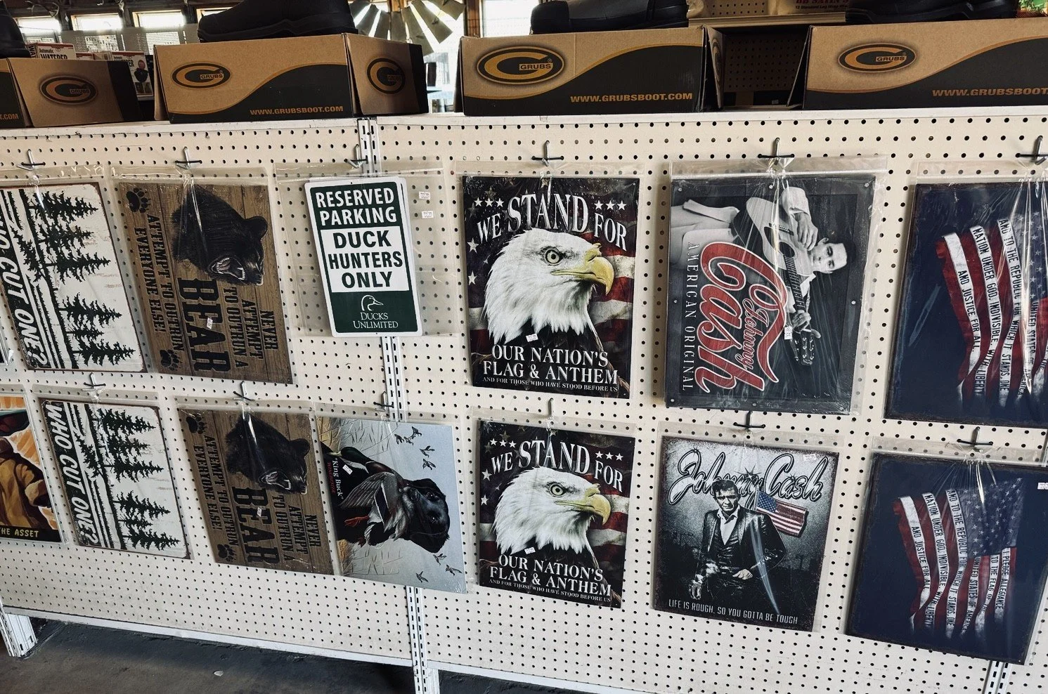 A display of patriotic and nature-themed metal signs and posters on a pegboard shelf. The signs include images of bears, eagles, the American flag, and messages supporting patriotism and hunting.