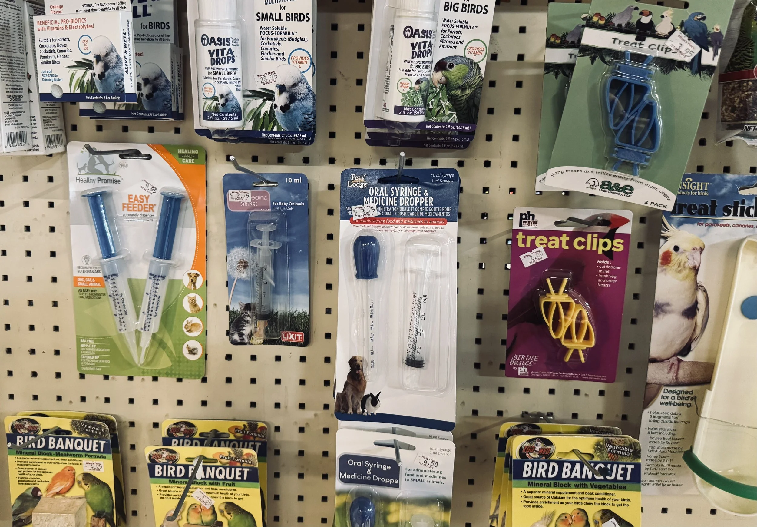 A display of bird care products on a store pegboard, including bird vitamins, oral syringes, treat clips, and bird food blocks with colorful bird illustrations and labels.