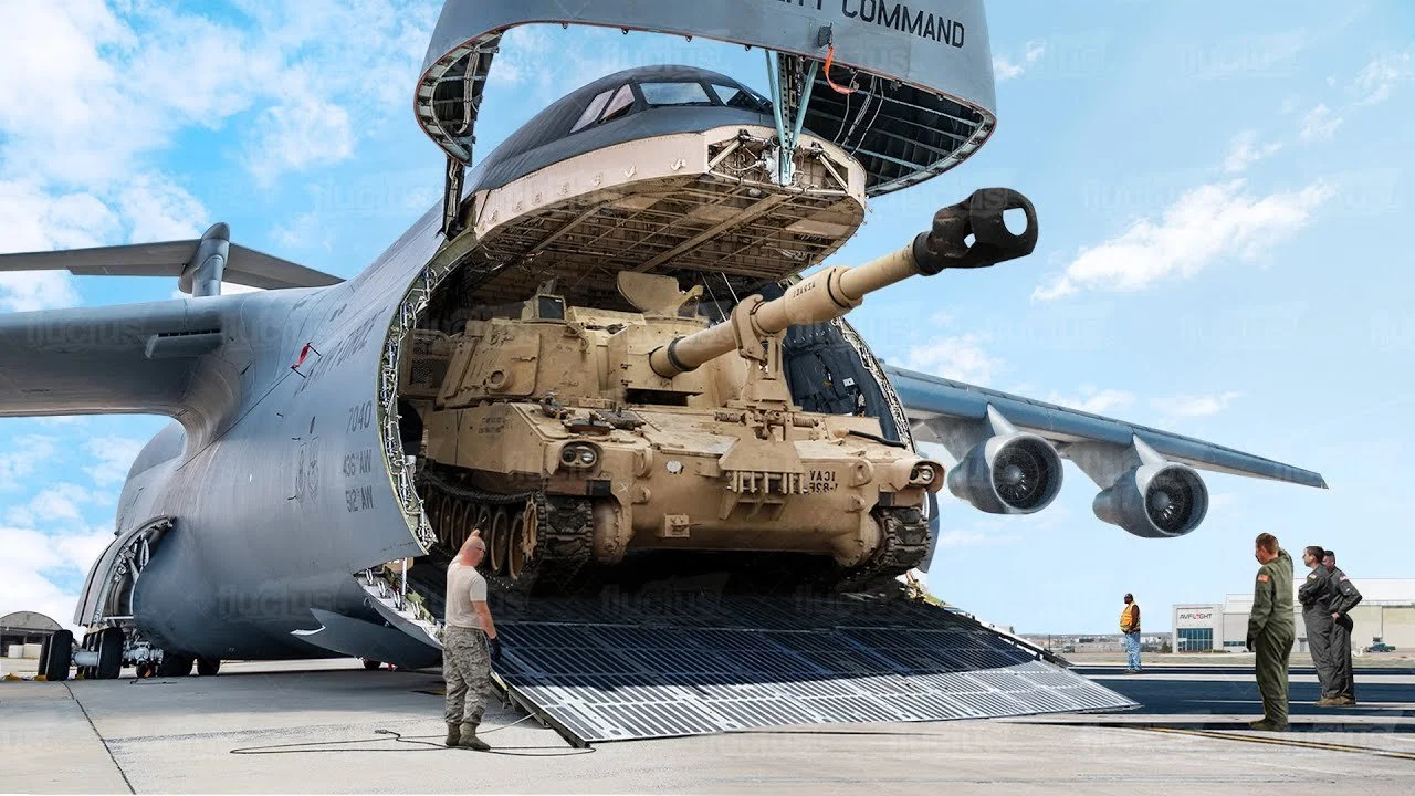 Military personnel loading a tan tank into a large cargo aircraft with a rear ramp during daytime, with a blue sky and clouds.