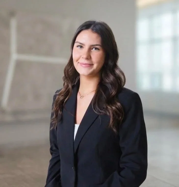 A young woman in a black blazer smiling, standing indoors with blurred background.