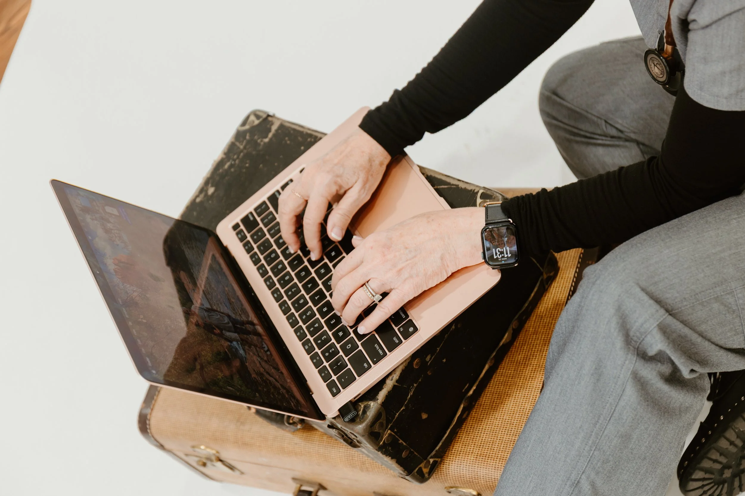 Person sitting on a vintage suitcase with a laptop on another suitcase, wearing a smartwatch and gray pants, against a plain white background.
