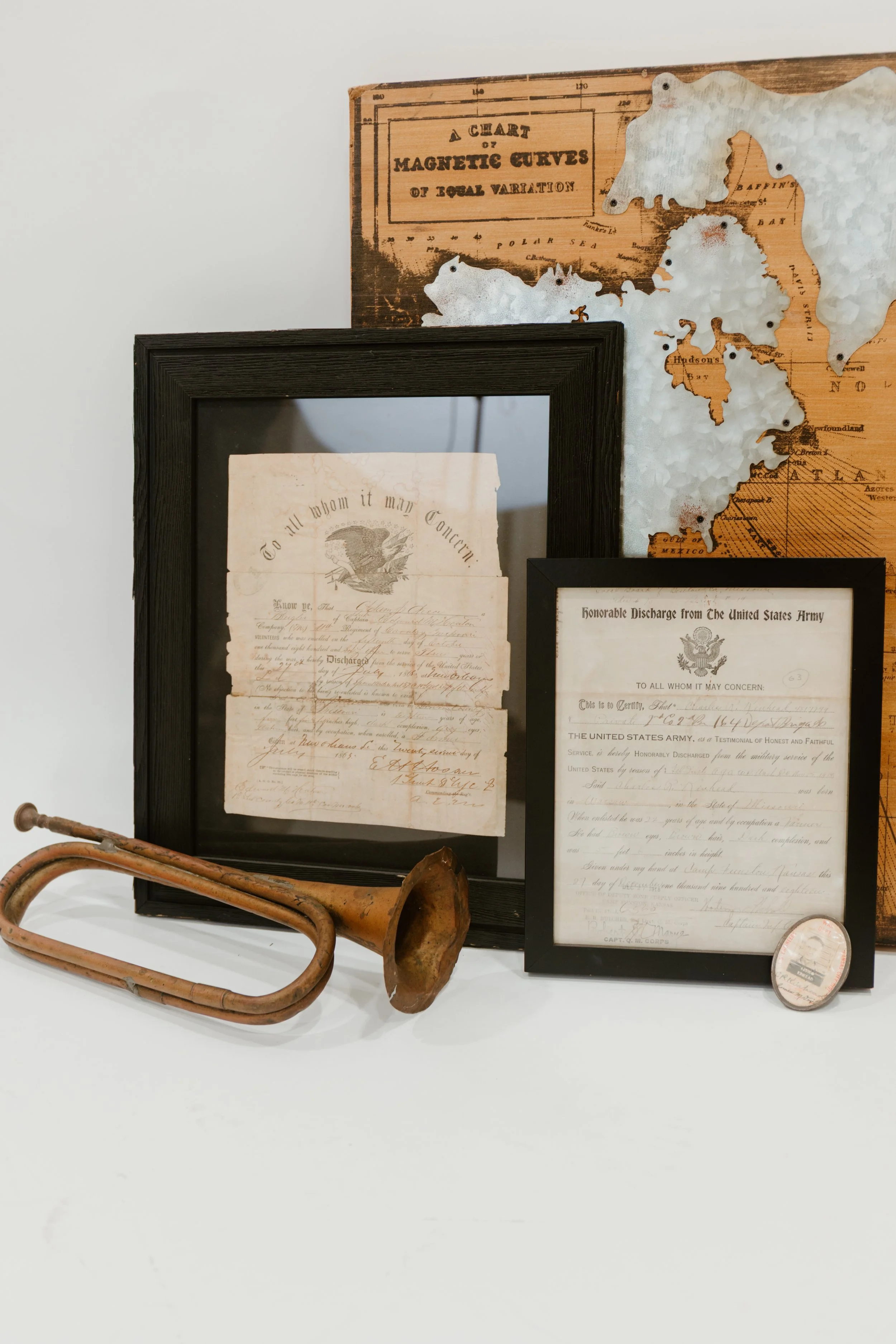 Vintage framed military discharges, ranging from the Civil War to Vietnam Era, leaning against a Vintage wooden/metal map, a brass Civil War Bugle lies in front of the Civil War discharge, with other historical items displayed on a white surface.