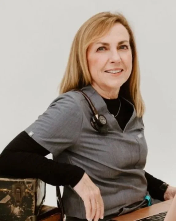 A smiling woman with shoulder-length blonde hair, wearing medical scrubs and a stethoscope, sitting with a laptop on her lap.