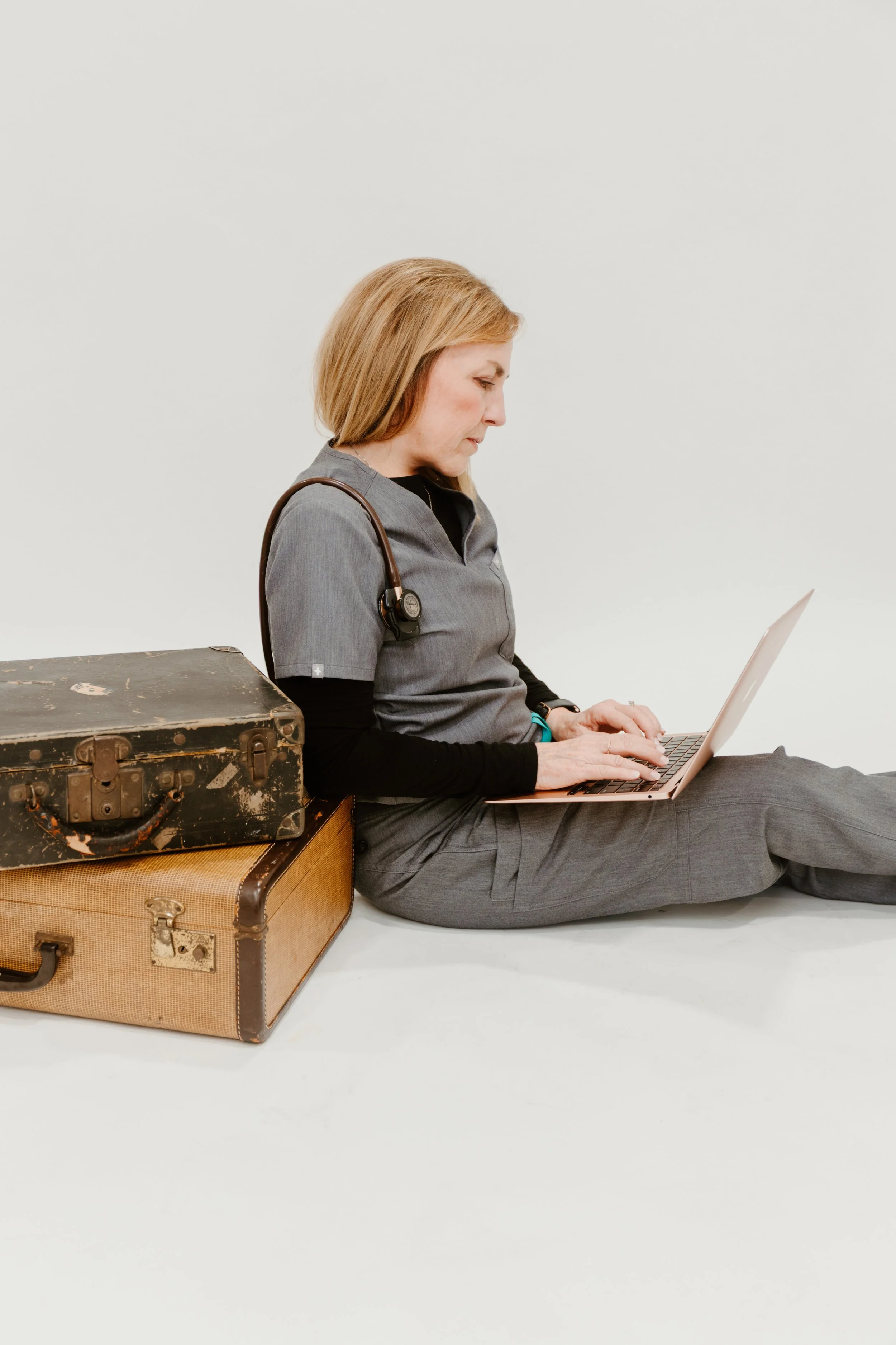 A woman with blonde hair wearing gray scrubs and a stethoscope is sitting on the floor, working on a laptop, while leaning against a couple of vintage suitcases.