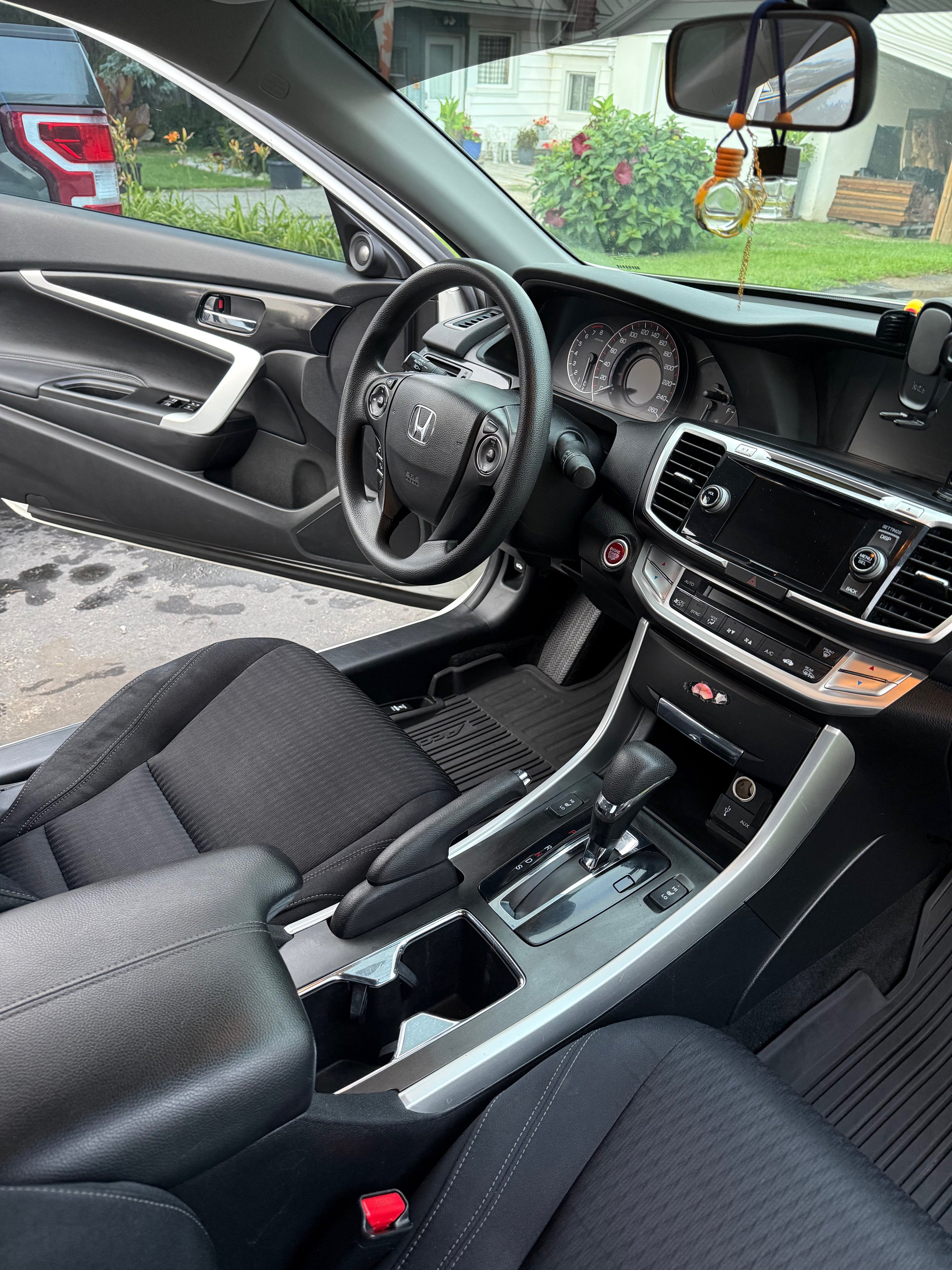 Interior of a car, showing the dashboard, steering wheel, center console with gear shift, and front seats with black upholstery. Through the windshield, a house, shrubbery, and a parked vehicle are visible outside.