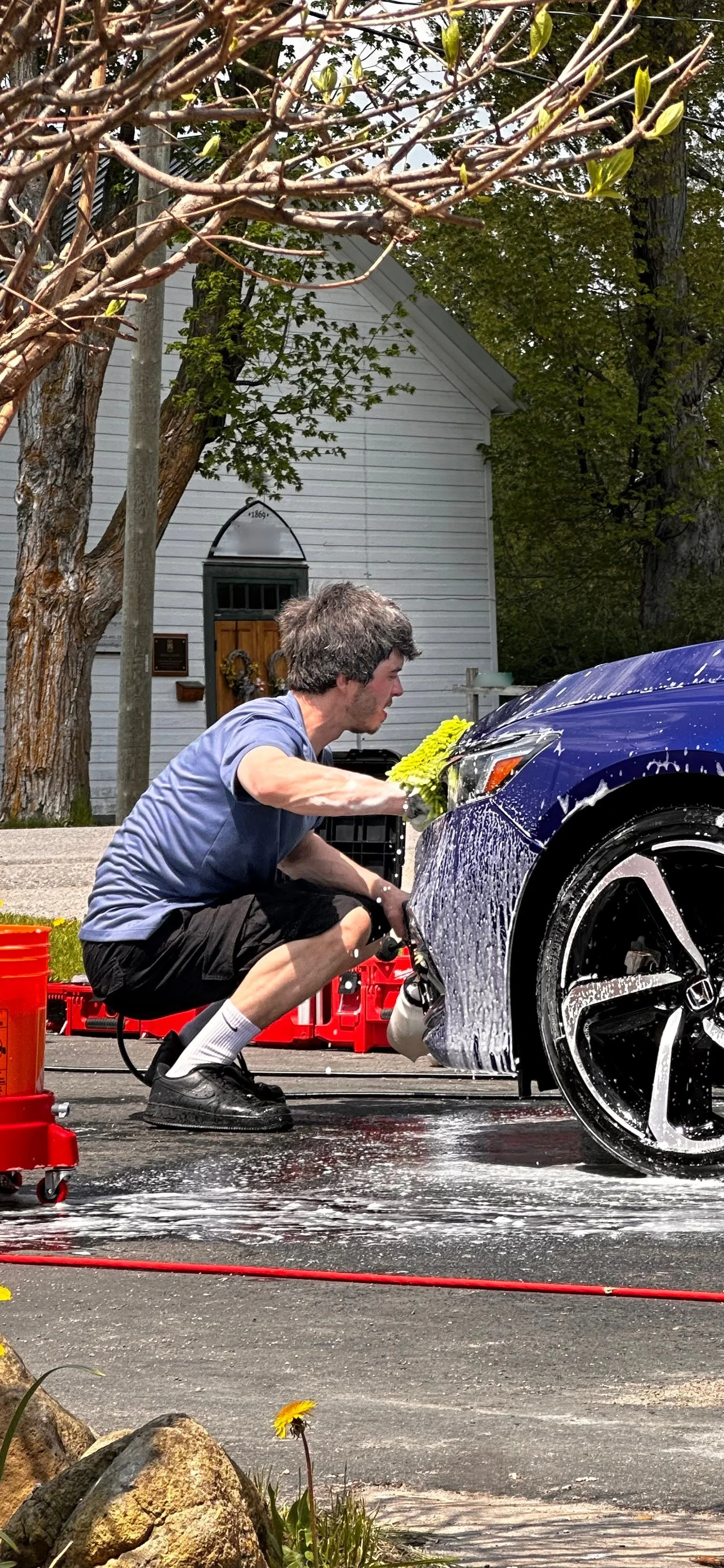 A man cleaning a blue car with a yellow sponge on a driveway outside a house.