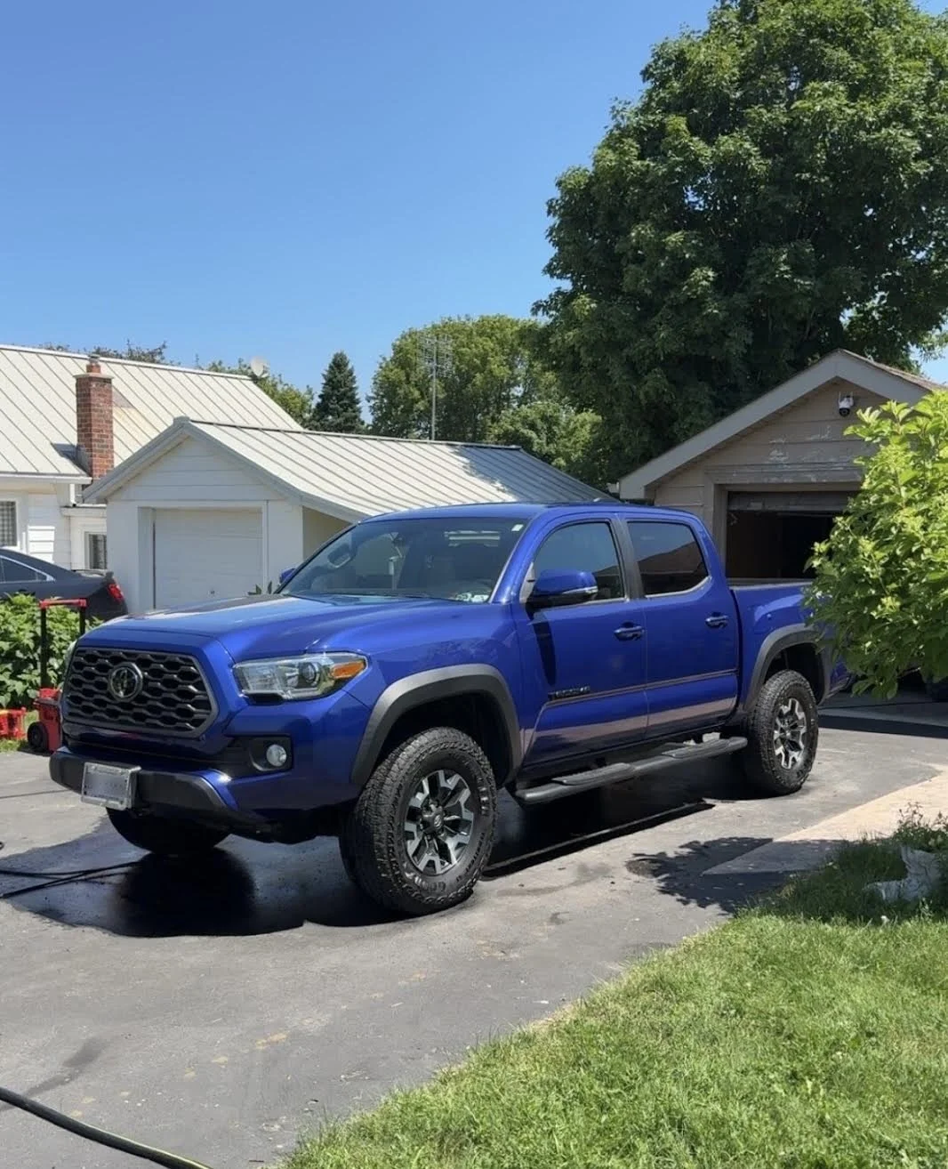 A blue Toyota pickup truck parked on a driveway in front of a garage, with a house and large trees in the background on a sunny day.