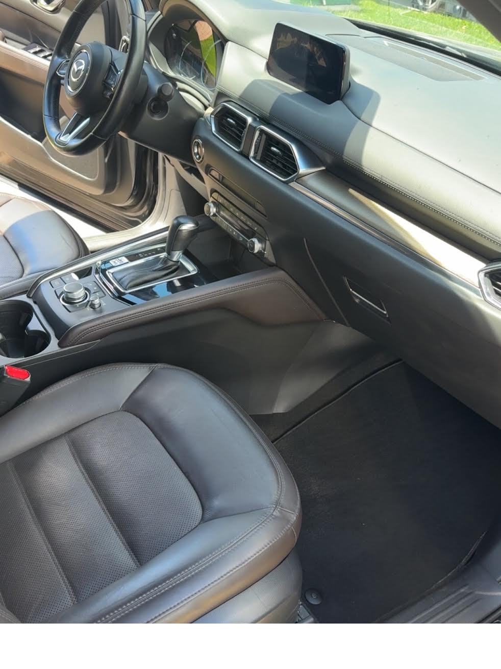 Interior of a modern vehicle showing the driver's seat, steering wheel with Mazda logo, gear shift, infotainment screen, and dashboard with air vents.