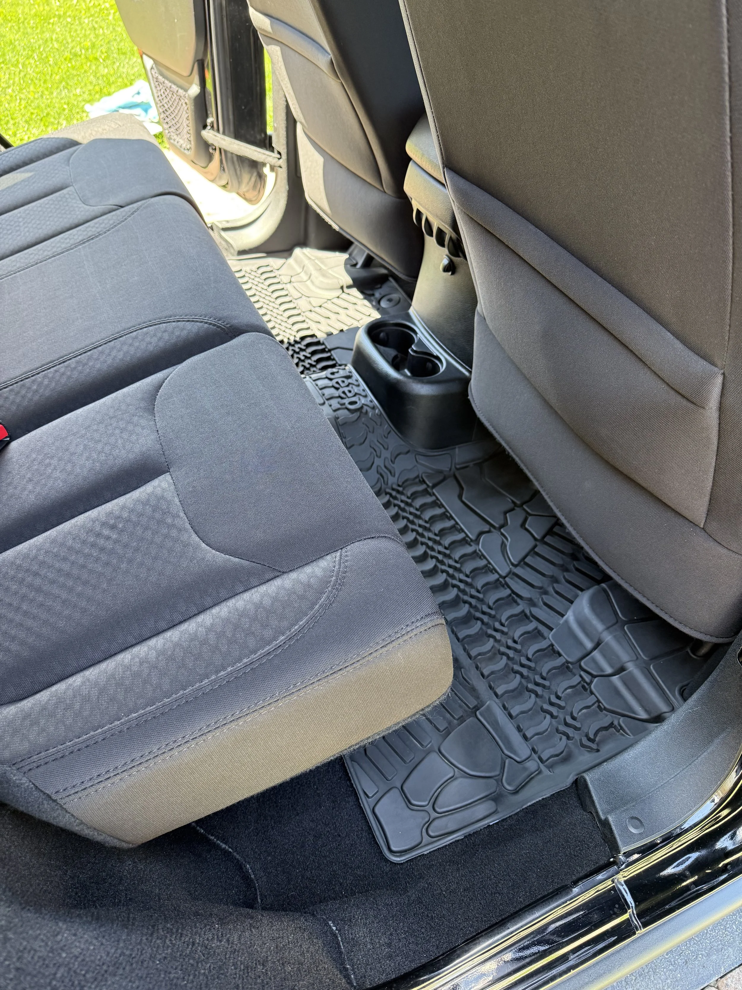The backseat area of a vehicle showing gray fabric seats, black rubber floor mats, and the back of two front seats with pockets. The vehicle door is open with a green grassy background visible outside.
