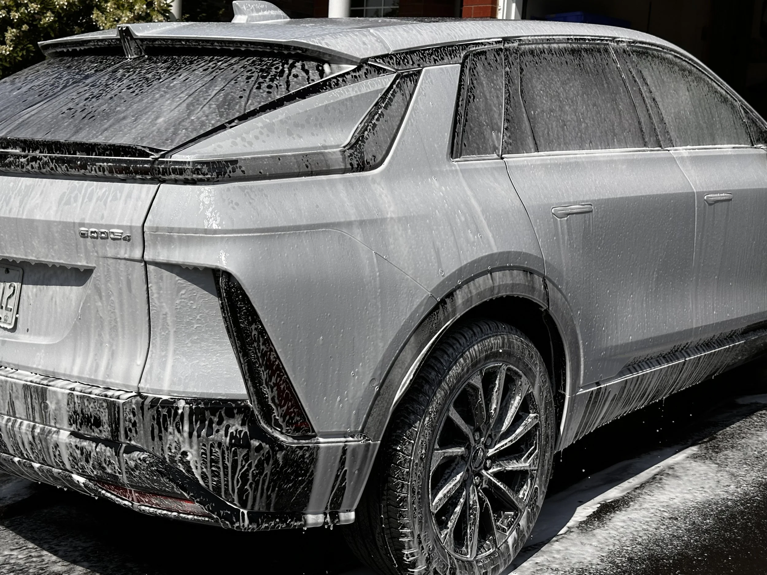 A silver Cadillac SUV covered in soap suds during a car wash, with water dripping down the body and foam on the windows, tires, and rear of the vehicle.