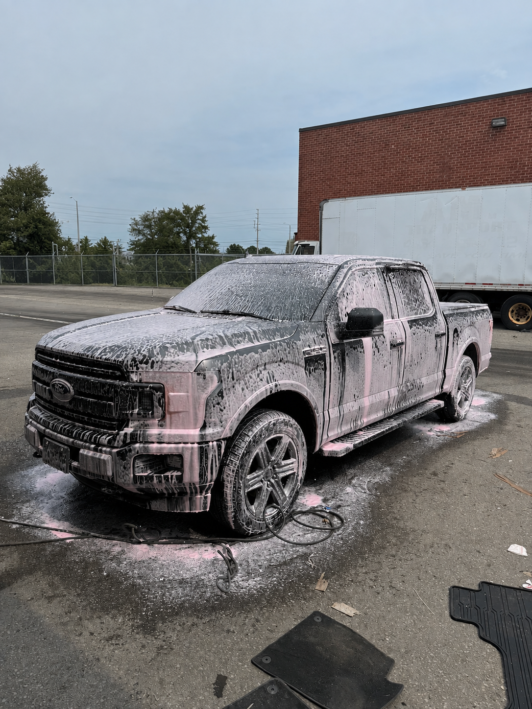 A black Ford pickup truck covered in pink foam and soap during a wash, with soap suds dripping down the sides, in a parking lot.