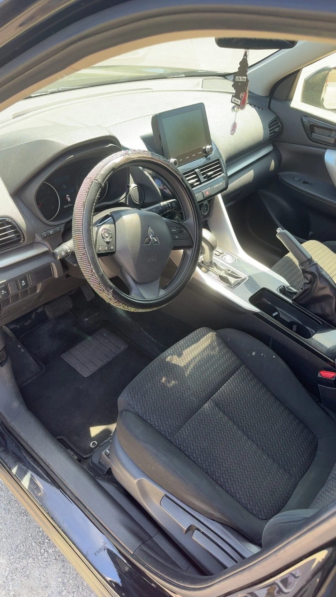 Inside view of a car interior, showing the steering wheel, dashboard, central console, gear shift, and front seats. The dashboard features a touchscreen display and hanging air freshener.