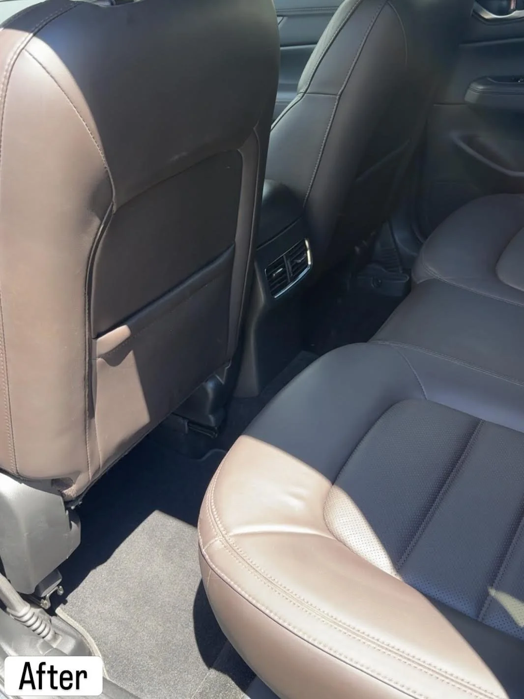 The backseat of a car showing two black leather seats, an air vent, and a floor mat.