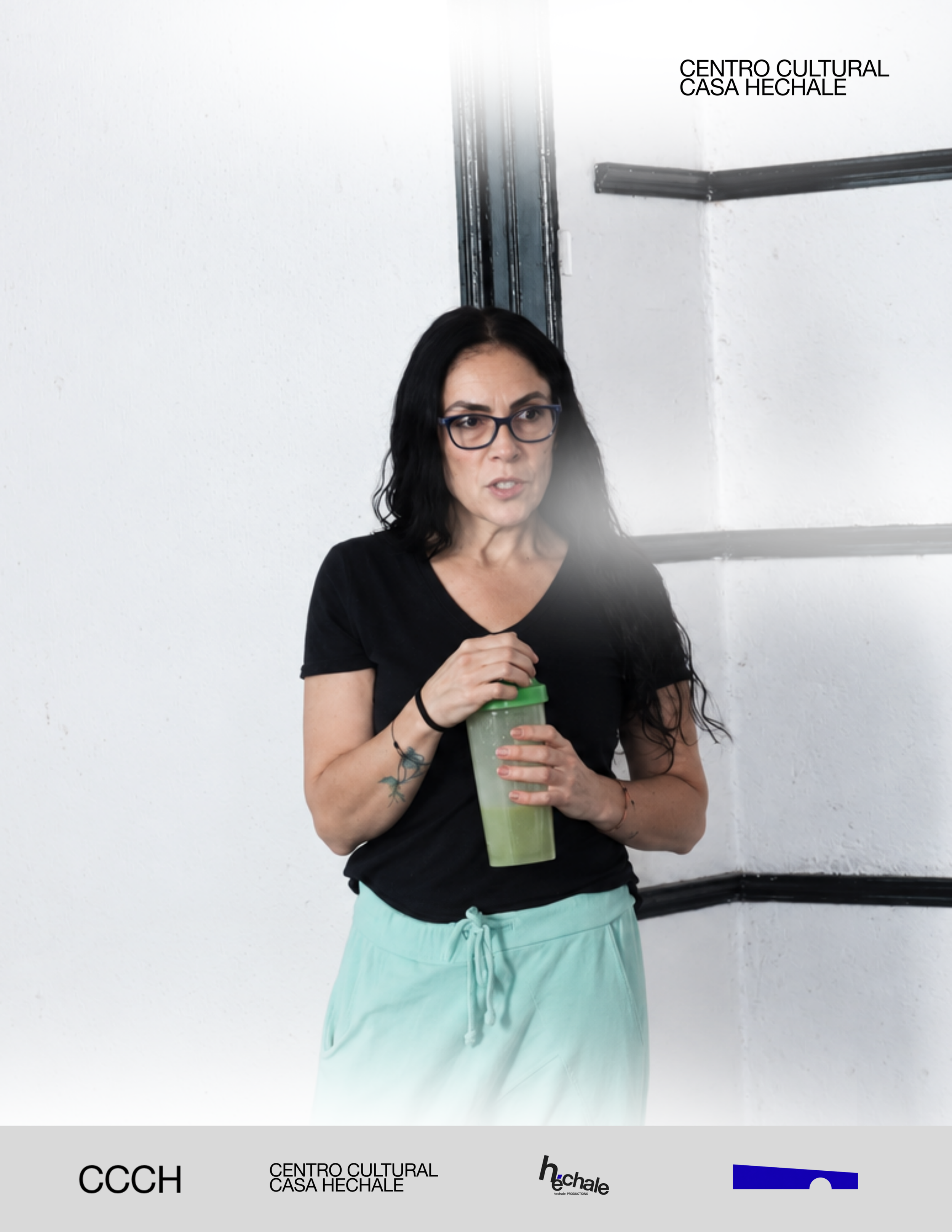 A woman with black hair and glasses, wearing a black shirt and light blue shorts, holding a green reusable tumbler, standing against a white wall near an industrial elevator shaft. awaken your artist, casa hechale