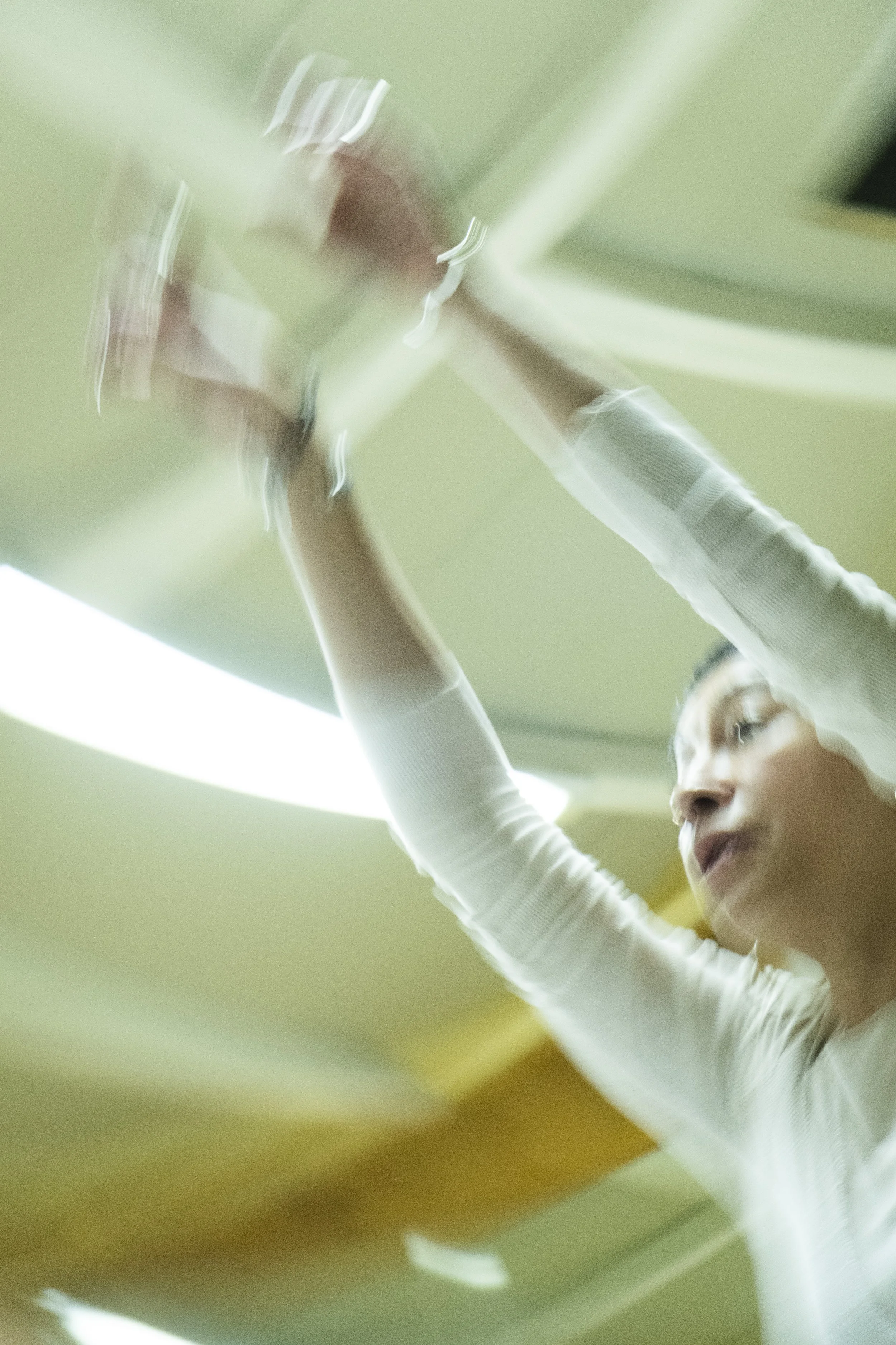 A woman with glasses and a white long-sleeve shirt reaching upward, with her arm extended, in an indoor setting with a green ceiling.
awaken your artist, casa hechale