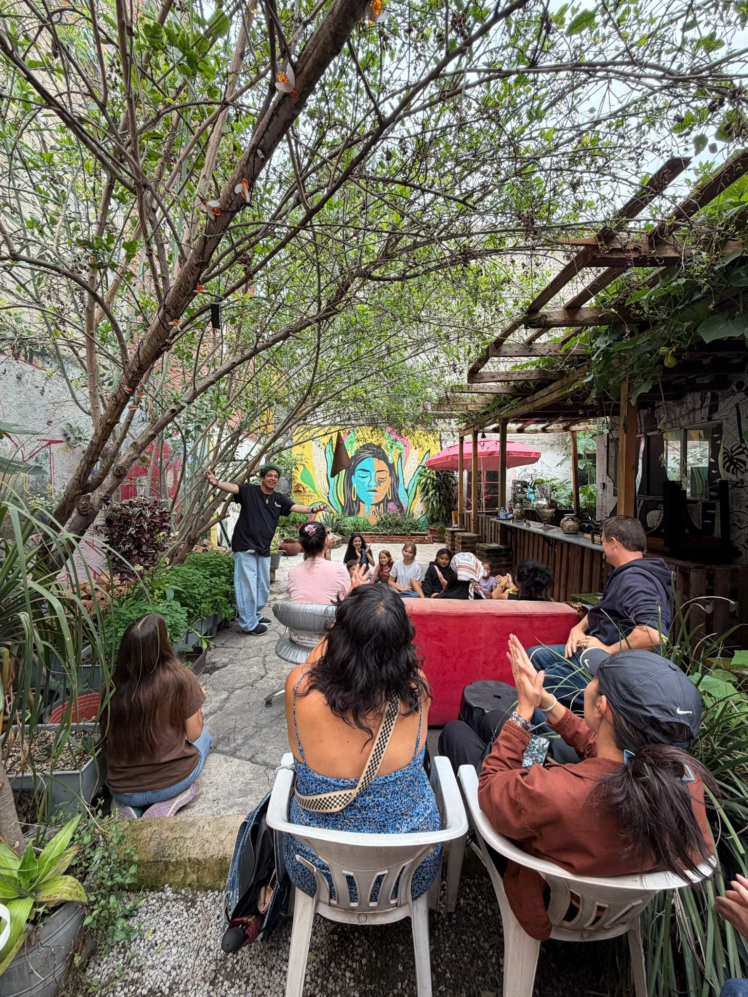 People gathered in an outdoor garden space, sitting on chairs and a bench, listening to a speaker standing near a colorful mural of a woman's face with closed eyes and flowers. There are plants, trees, and a small patio with a red umbrella in the bac