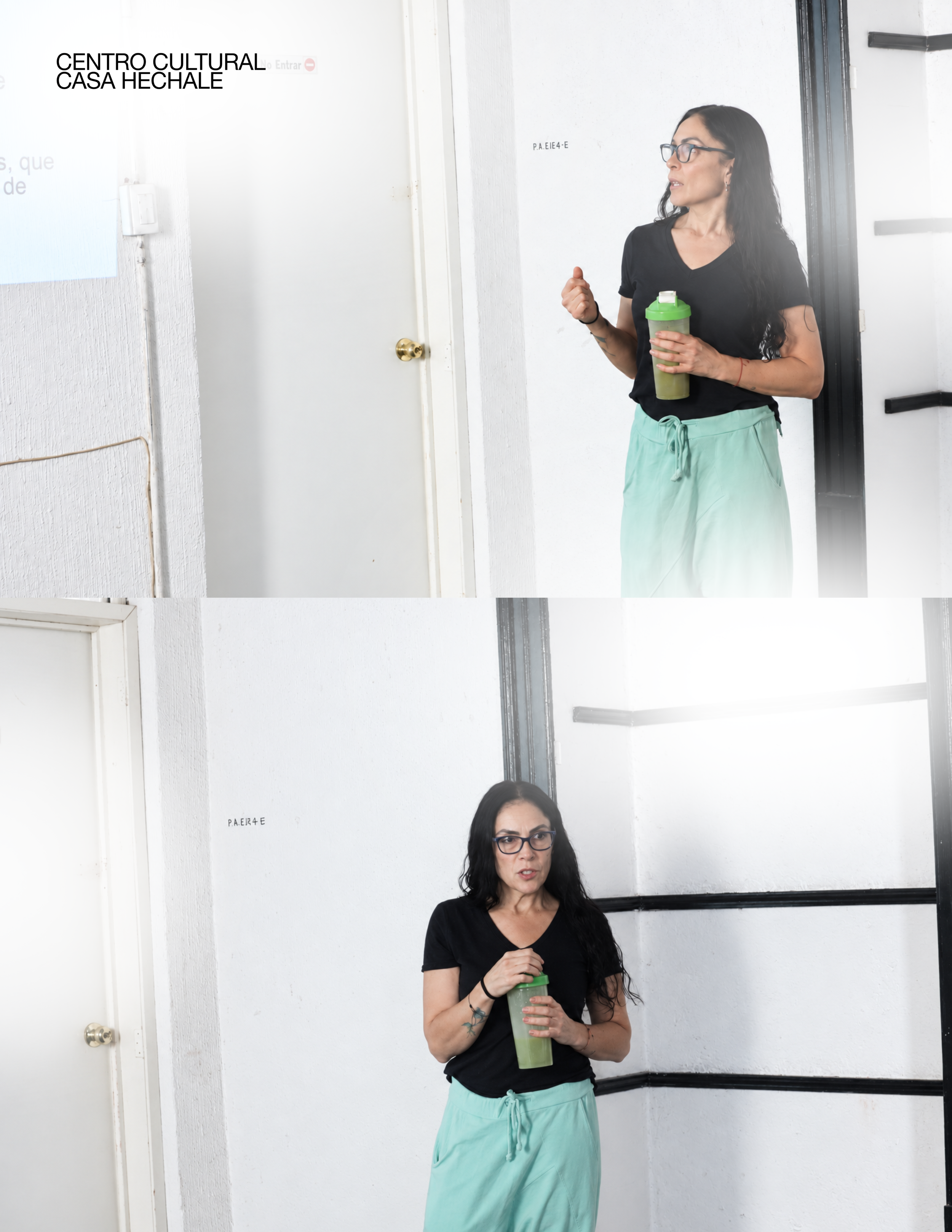 A woman with dark hair, glasses, and tattoos on her arm, standing indoors holding a green drink container, wearing a black t-shirt and light green sweatpants, in front of two white walls with text and a black frame. awaken your artist, casa hechale