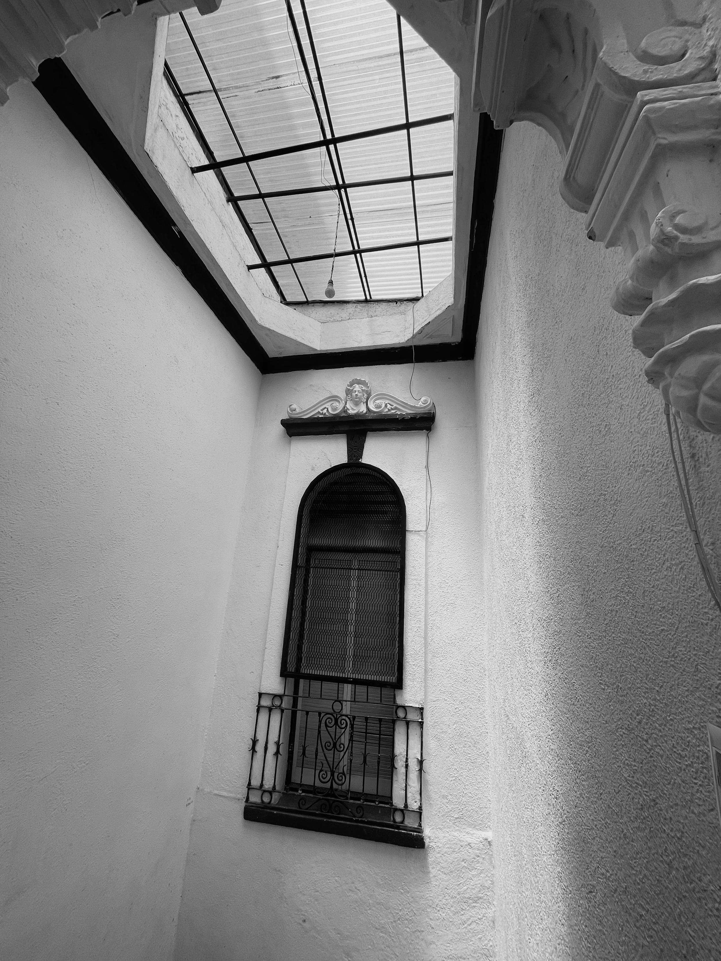 A view of a small enclosed courtyard with a decorative balcony, a window with wrought iron bars, and a skylight with grid framing, all painted in white.