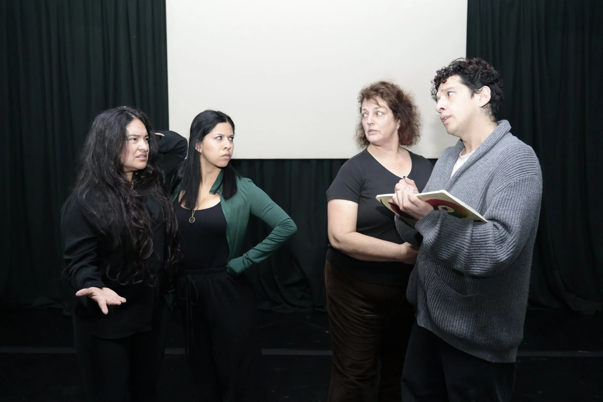 A group of four people in conversation, standing in front of a large white screen and black curtains, with some holding notebooks.