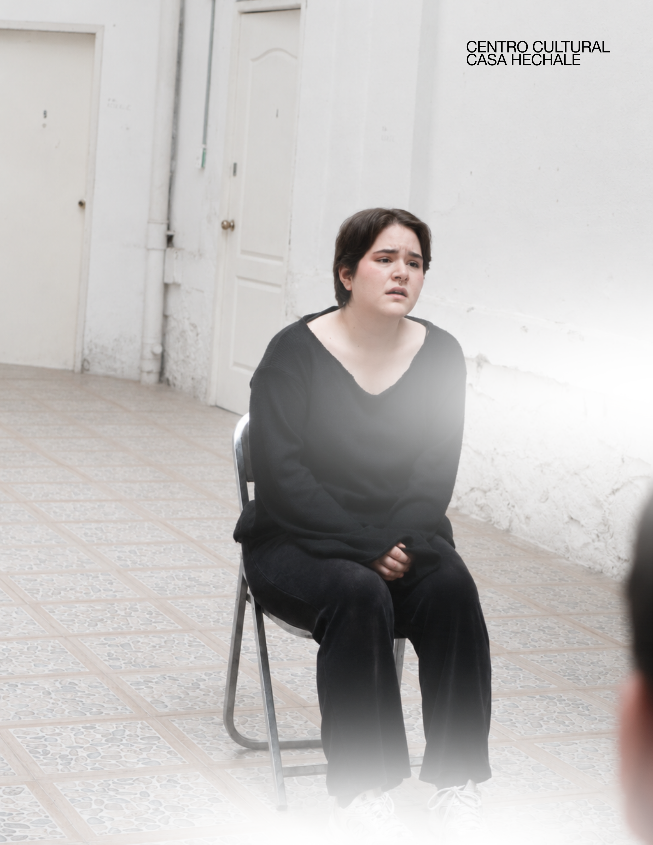 A young woman with short dark hair sitting on a metal chair, wearing a black top and pants, with a distressed facial expression, indoors against white walls and floors, with a sign saying 'Centro Cultural Casa Hechale' on the wall.