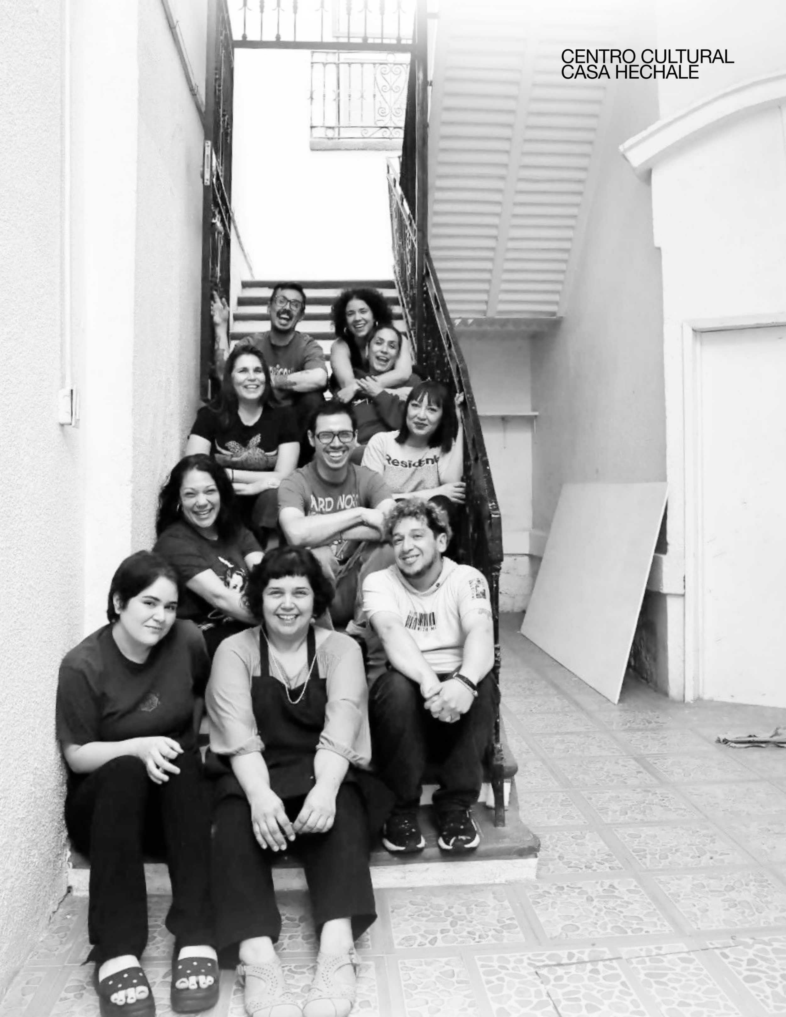 A group of eleven people, mostly young adults, sitting and standing on a staircase inside a building, smiling and posing for the photo. The staircase has a decorative metal railing, and there's a sign on the wall that reads "Centro Cultural Casa Hech