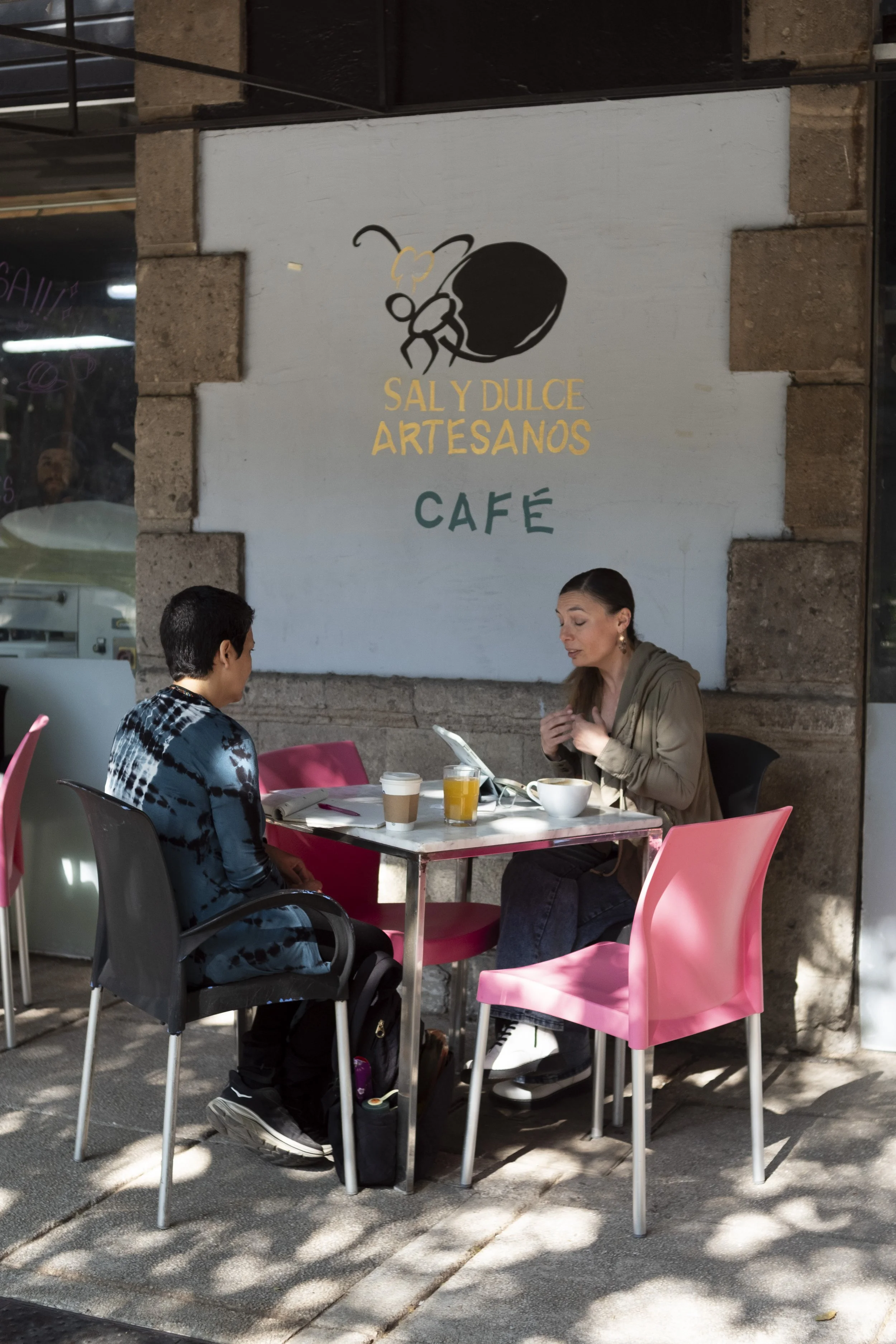 Two women having a conversation at an outdoor café table. The woman on the left has short black hair and is wearing a tie-dye shirt, while the woman on the right has long brown hair and is wearing a beige jacket. Their table has drinks and a bowl.