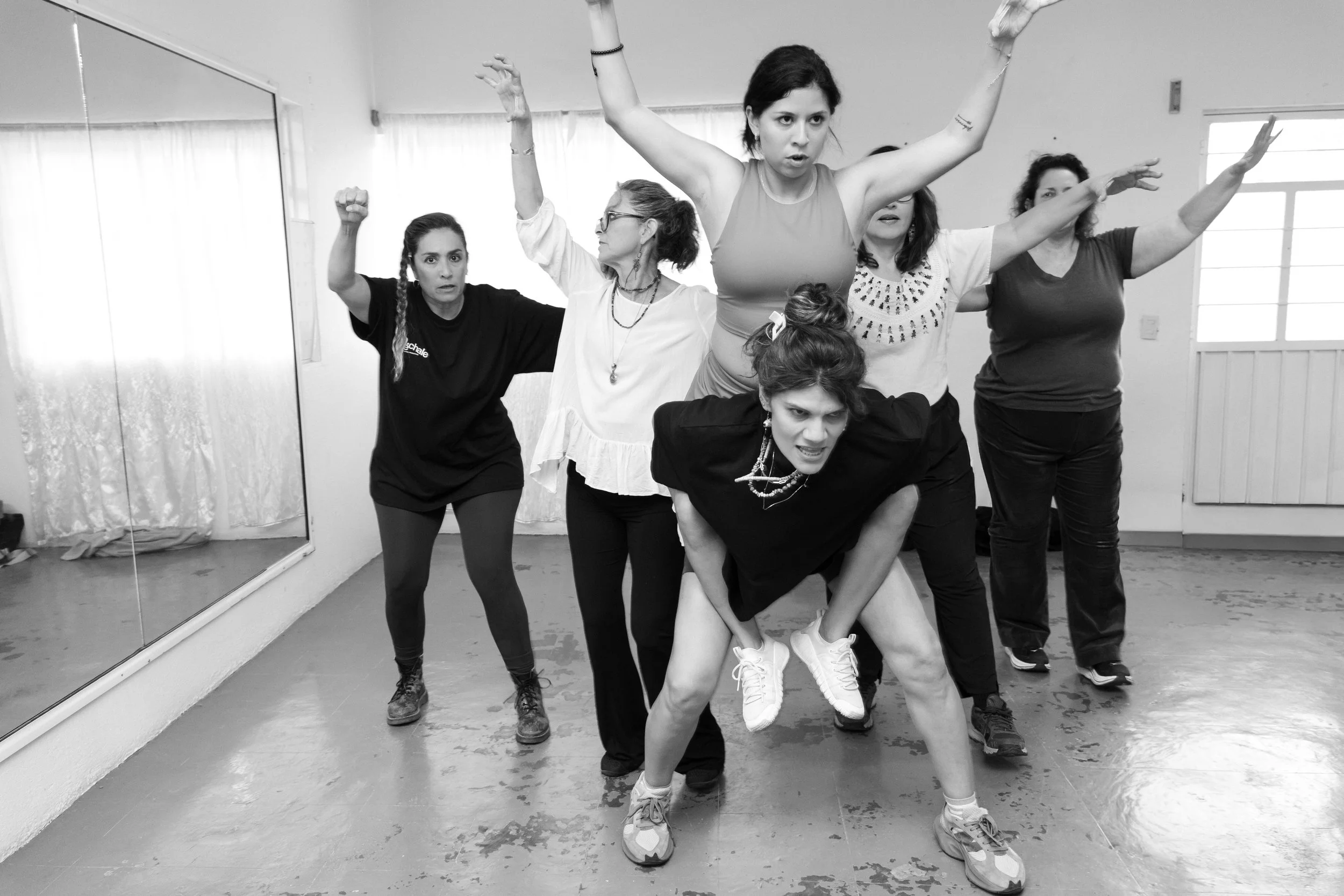 A group of women in a dance studio, participating in a dance or theatrical performance. Four women are standing in a row, with one woman crouching in front, all with expressive poses, and one woman in the background. They appear to be engaged in a co