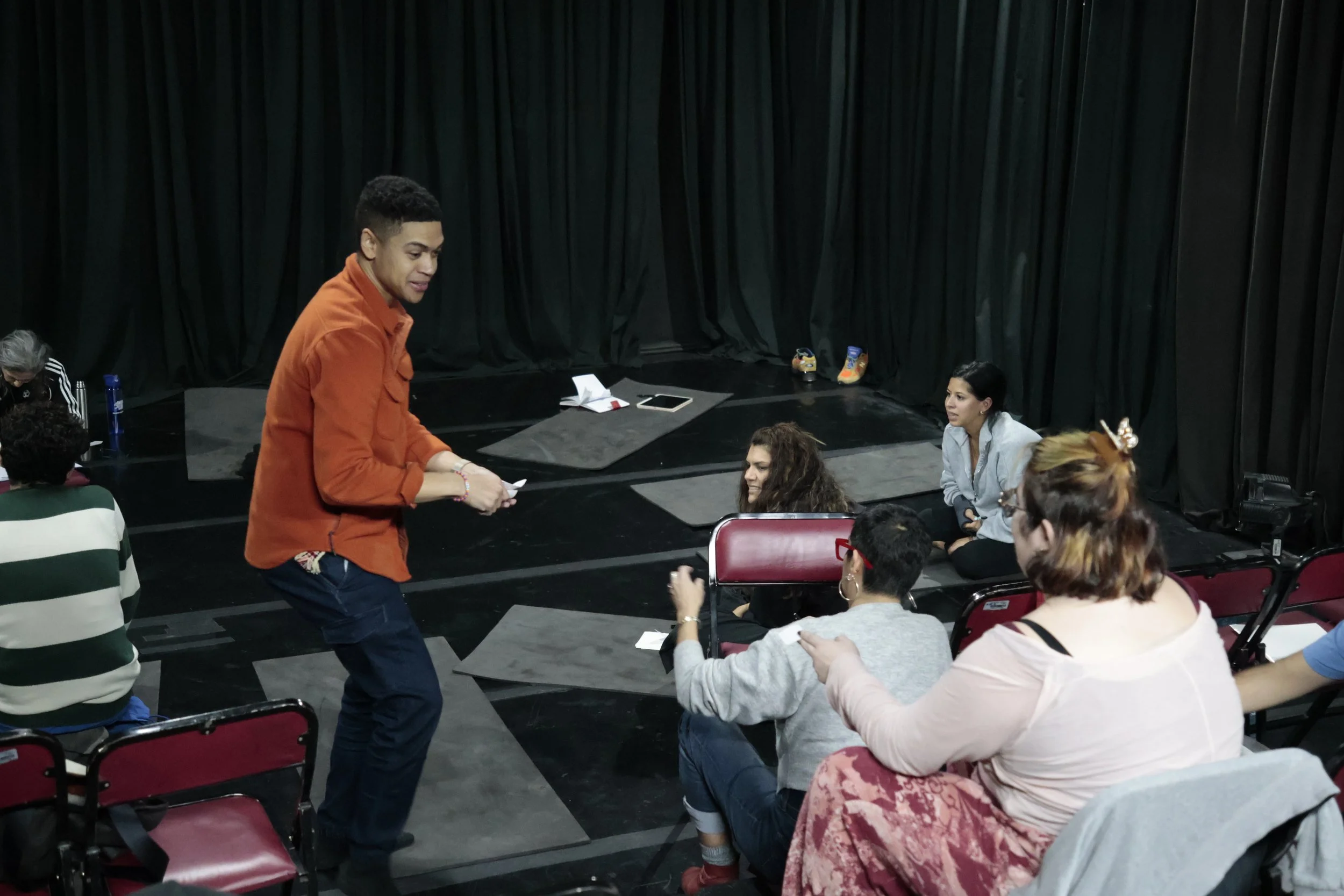 A group of diverse people seated in a theater or auditorium, with a young man in an orange shirt standing and talking to a woman in a gray jacket. Black curtains are in the background, and there are personal items on the stage.
