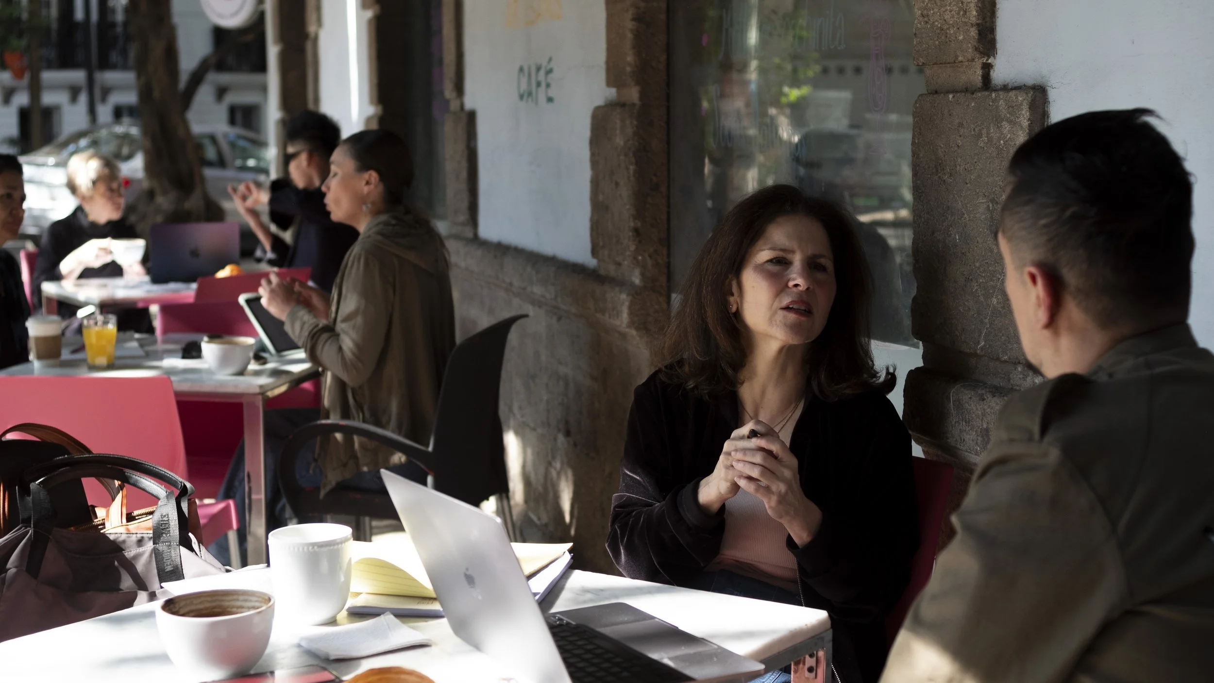 People sitting at tables inside a cafe, engaged in conversation, with coffee cups and laptops on the tables, and a large window letting in natural light.