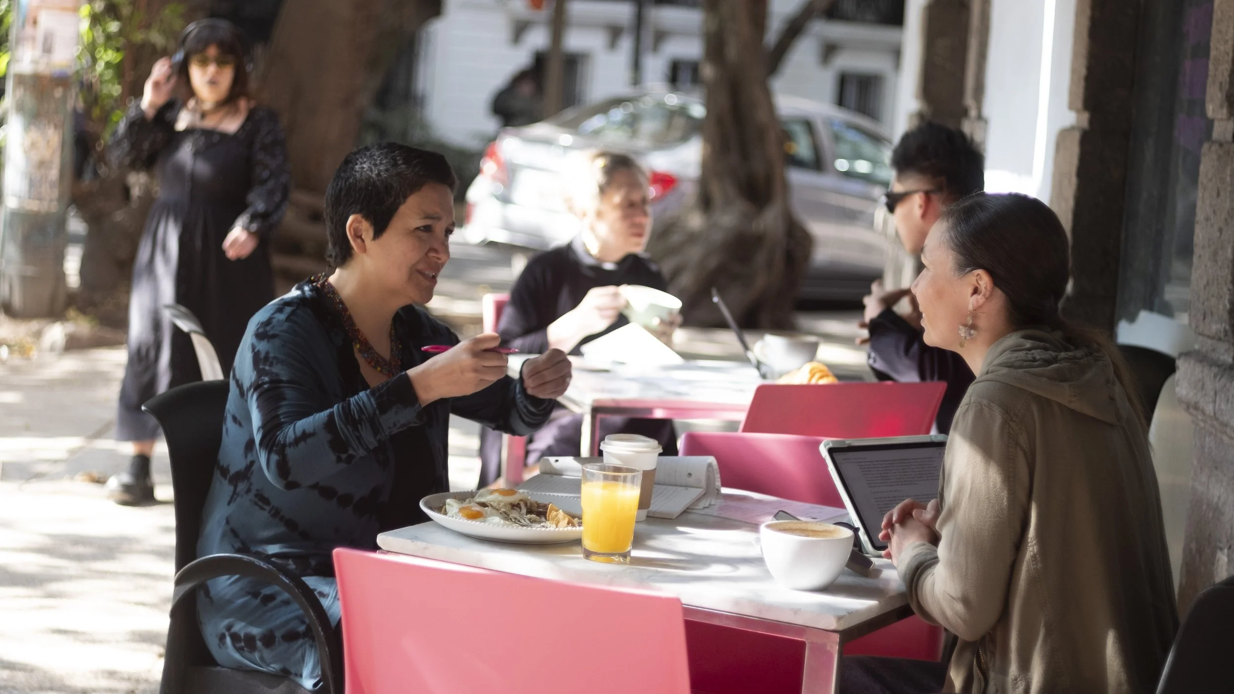 A group of people having breakfast outdoors at a cafe table, with women eating and talking, including one woman with a laptop and another with a tablet.