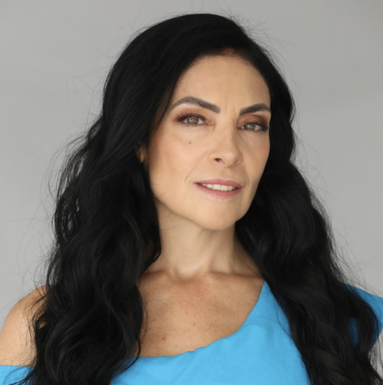 A woman with long black hair styled in waves, wearing a blue top, looking at the camera with a slight smile against a gray background.