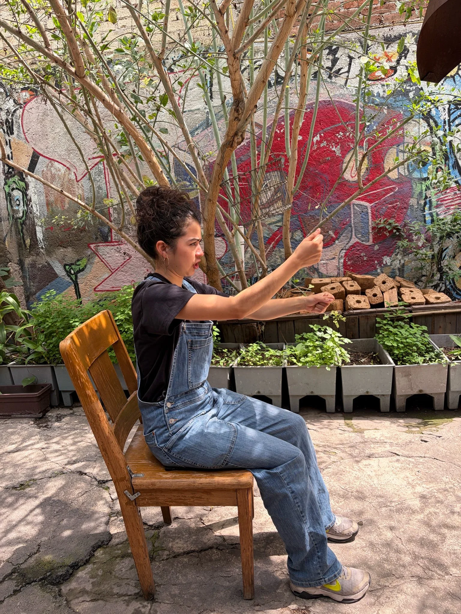 A woman with curly hair and light skin is sitting on a wooden chair outdoors, reaching with her right hand. She is wearing a black t-shirt, denim overalls, and sneakers, with a graffiti-covered wall and plants behind her.