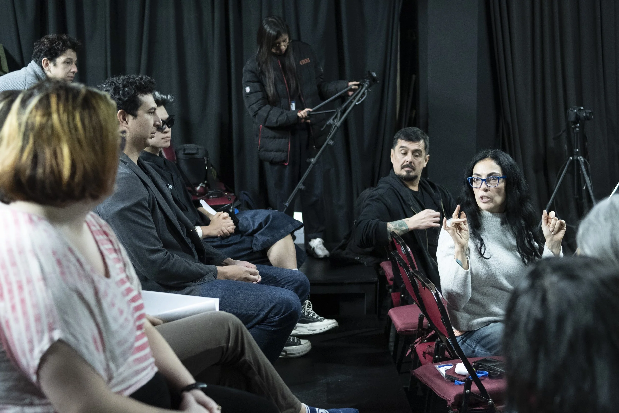 Group of people seated in a panel discussion or meeting, with one woman standing and speaking while others listen, in a room with black curtains and equipment. awaken your artist, casa hechale