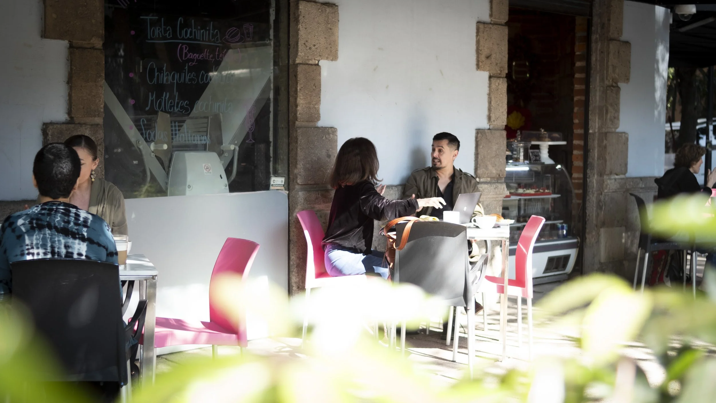 People sitting outdoors at a cafe, engaged in conversation, with a bakery display and a chalkboard menu in the background.